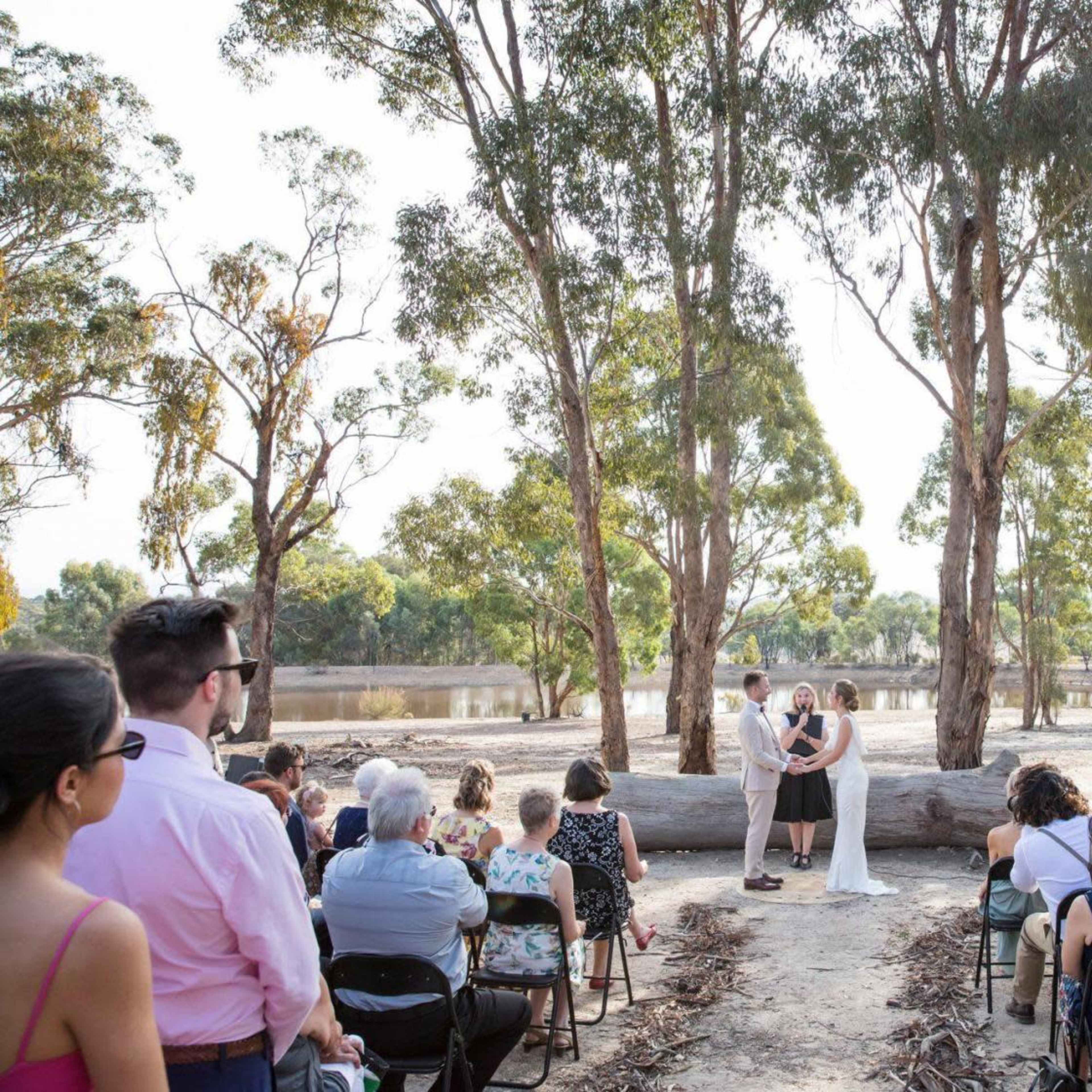 A couple stands in front of a gathering of guests under trees by a body of water during a wedding ceremony.