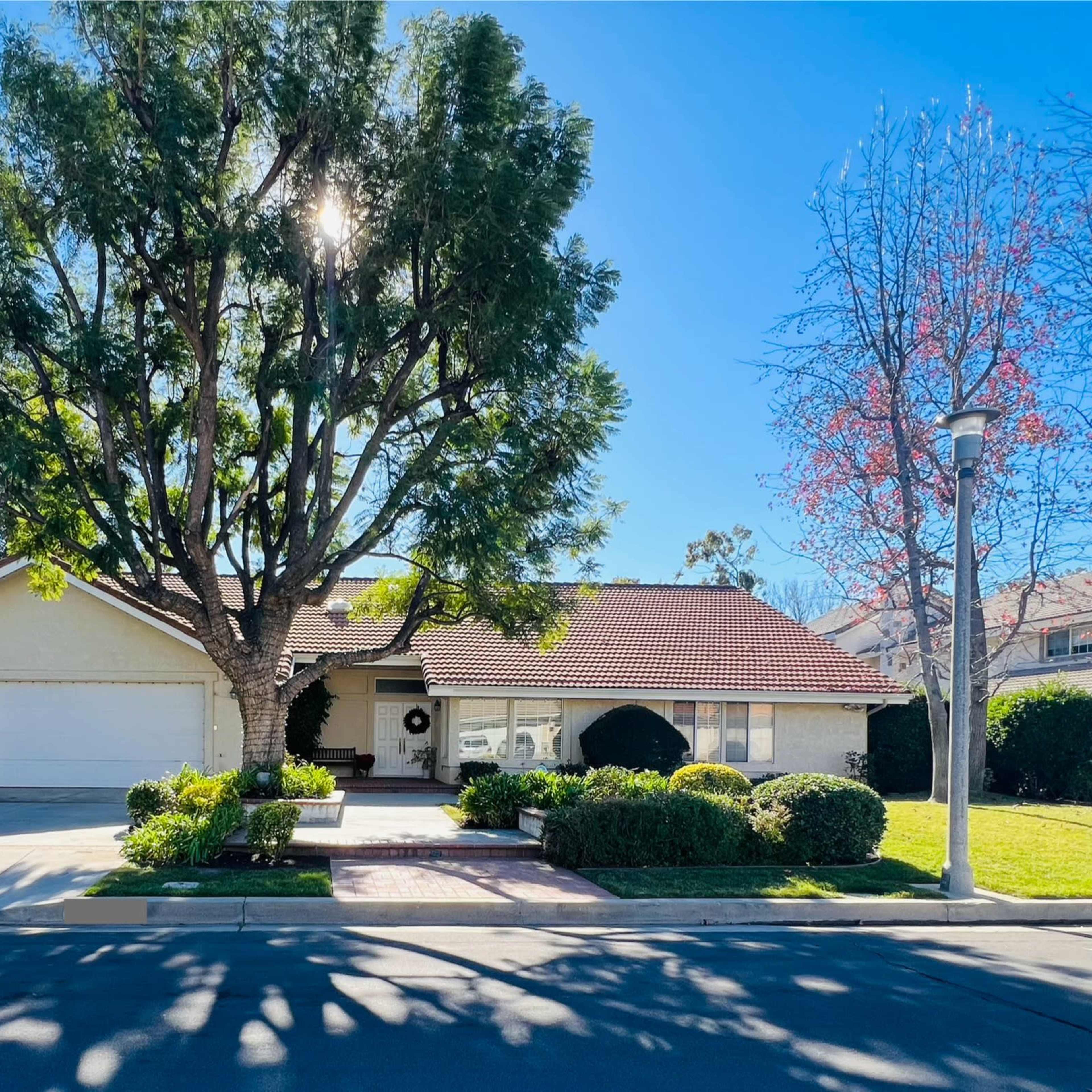 A single-story house with a red-tiled roof and two garage doors is surrounded by neatly trimmed shrubs and a large tree in the front yard.