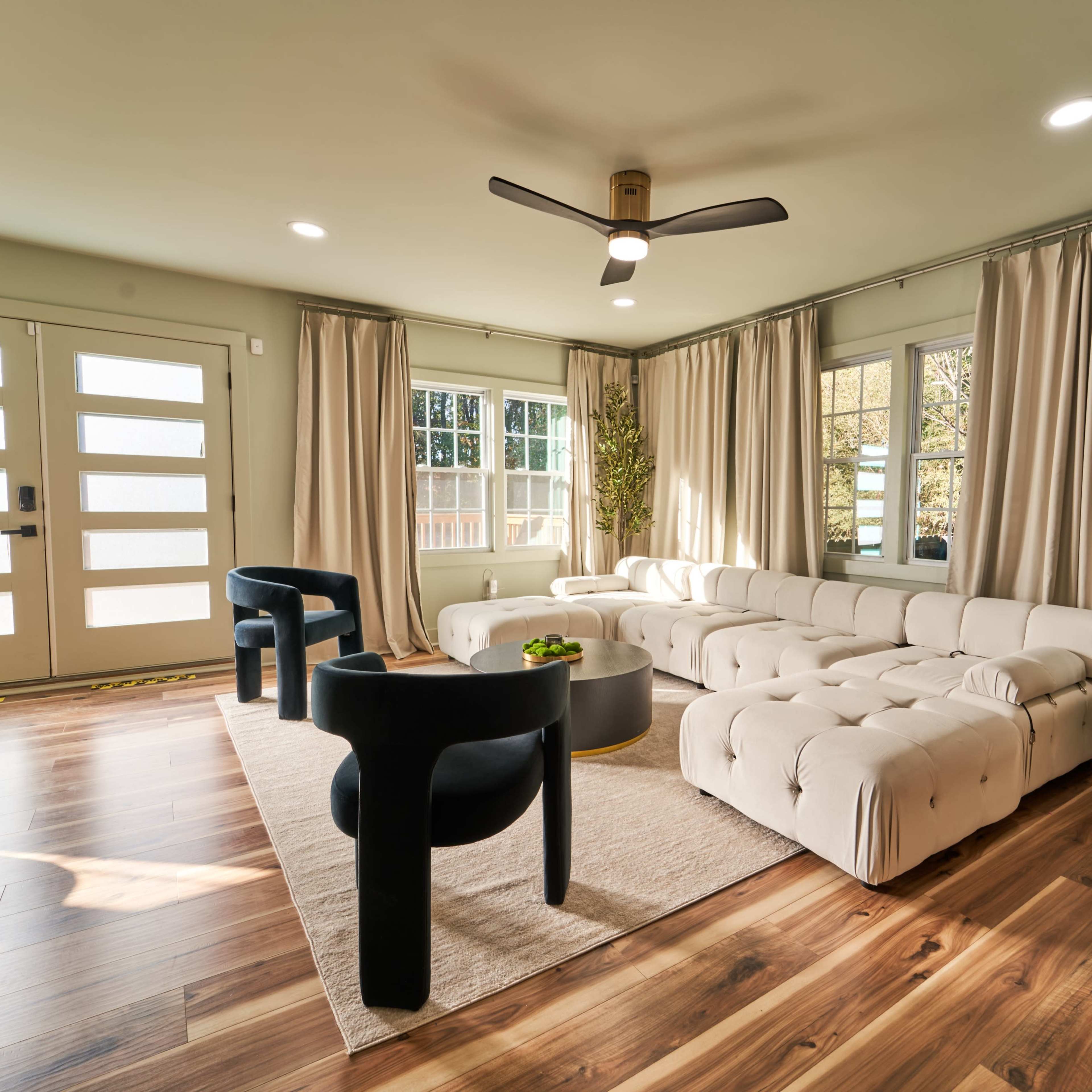 A modern living room features a light-colored sofa, black accent chairs, a coffee table, and a Christmas tree beside large windows.