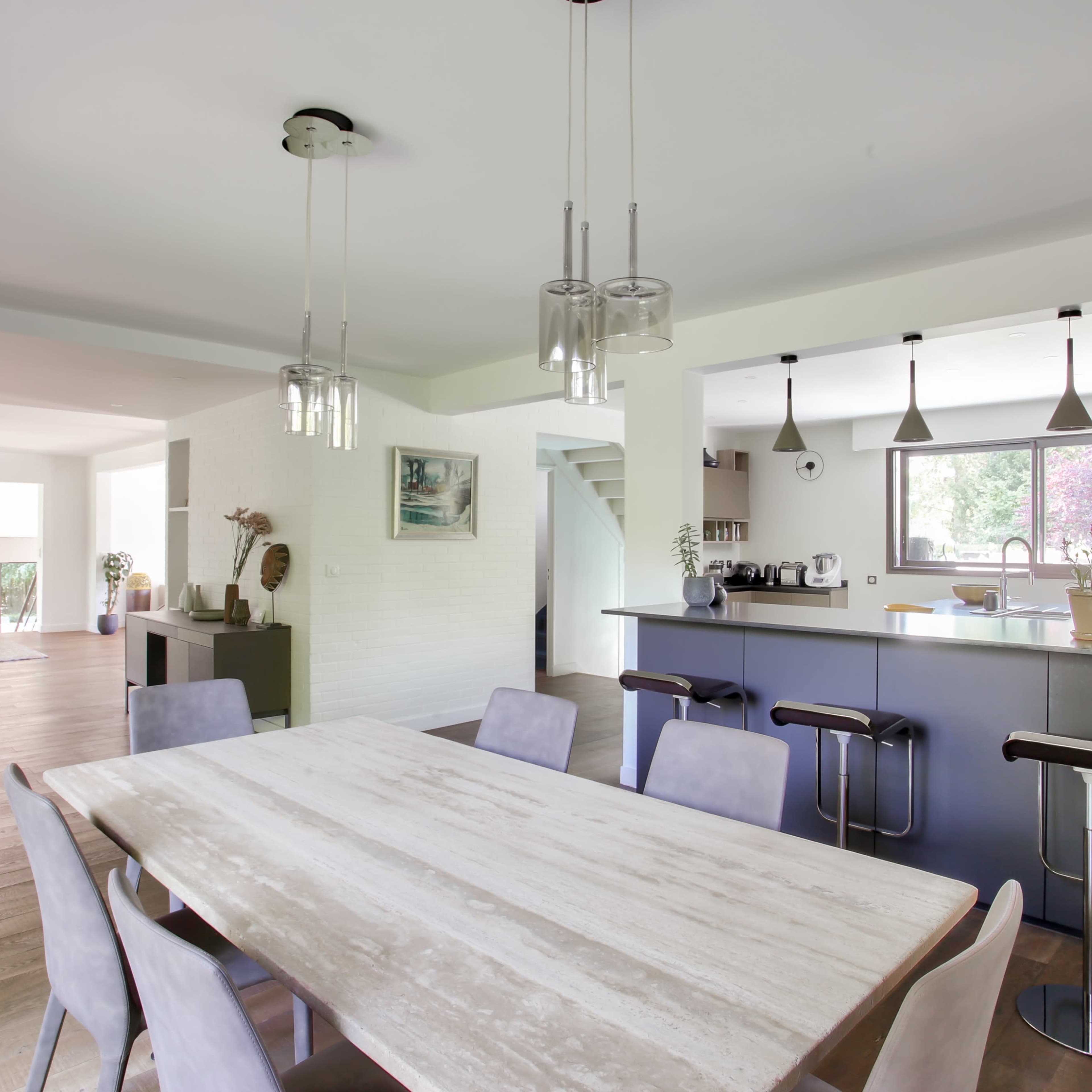 A modern open-concept living and dining area with a wooden dining table, bar stools at a kitchen counter, and large windows allowing natural light.