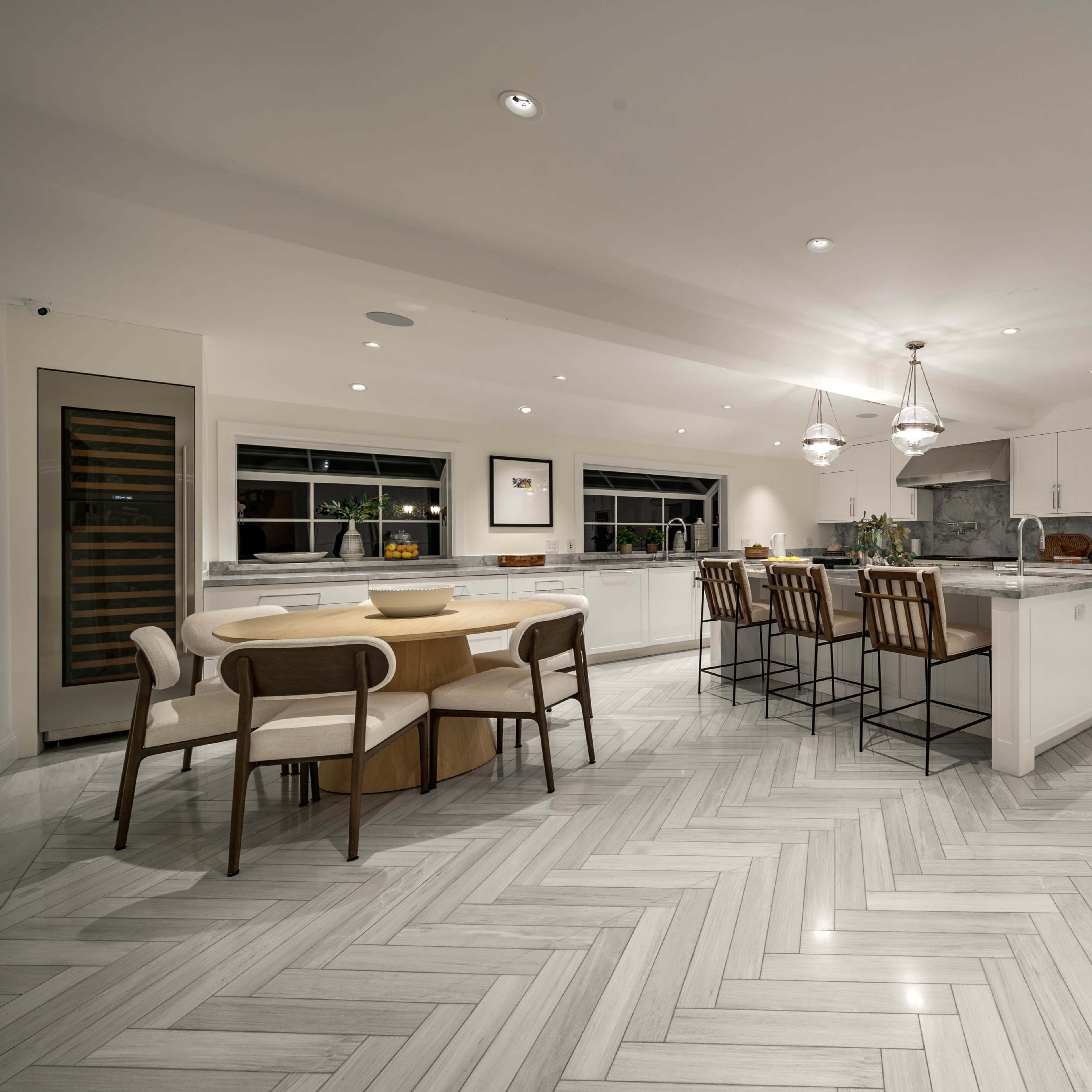 A modern kitchen with a large island, bar seating, and a dining table surrounded by chairs, all featuring polished marble flooring arranged in a herringbone pattern.