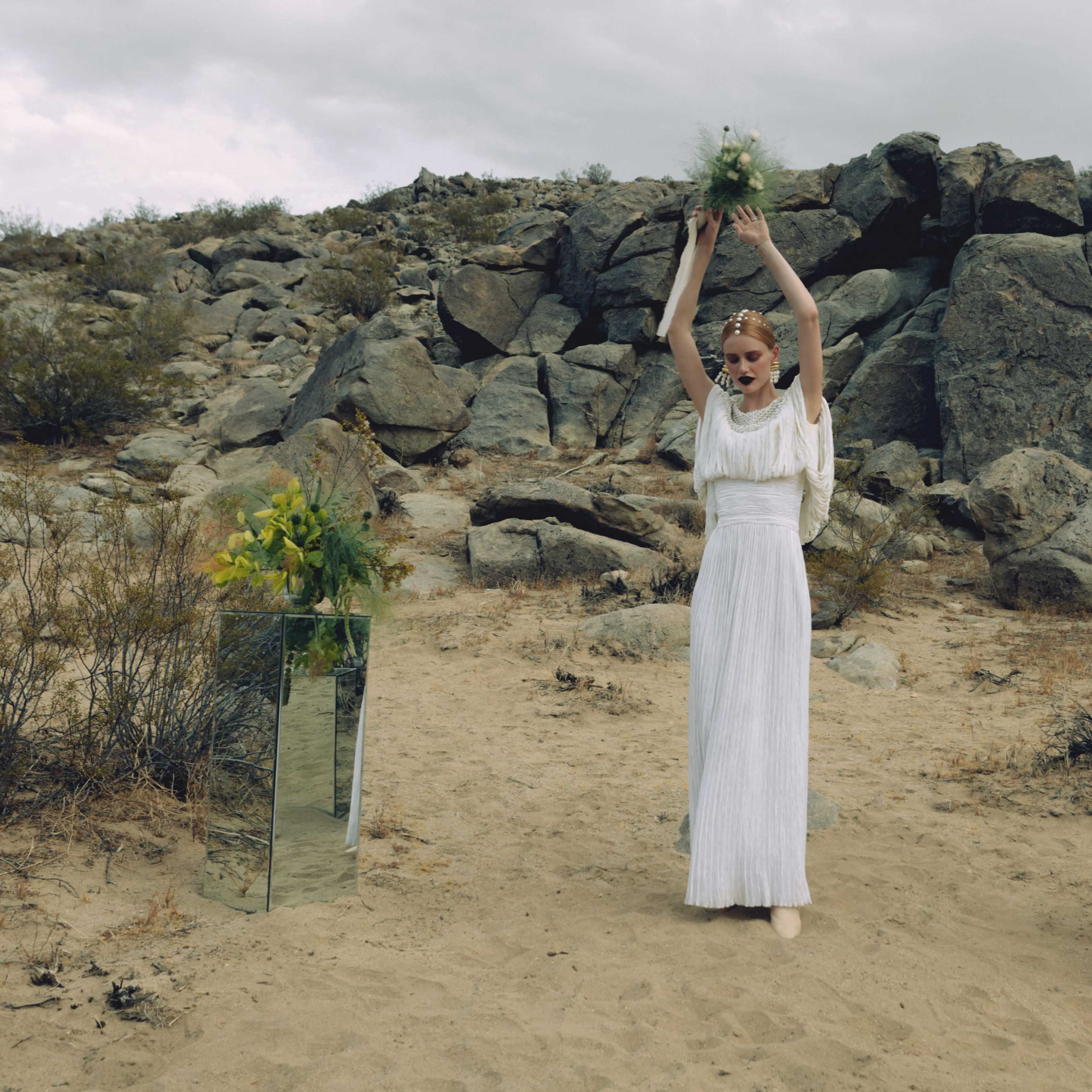 A woman in a white dress holds a bouquet of greenery above her head in a rocky desert landscape.
