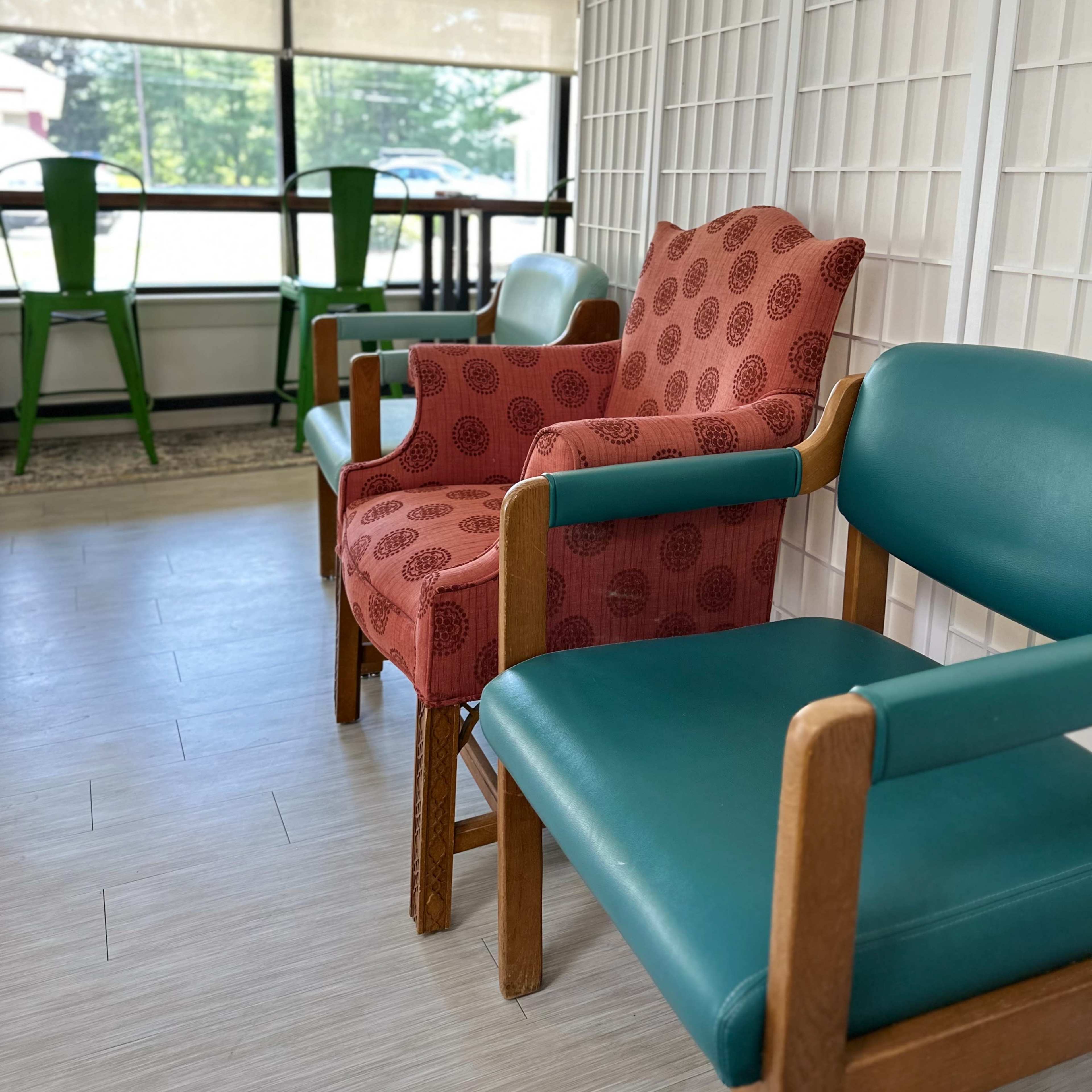 A waiting area features two wooden chairs, one with a patterned red cushion and the other with a teal seat, beside a large window with natural light.