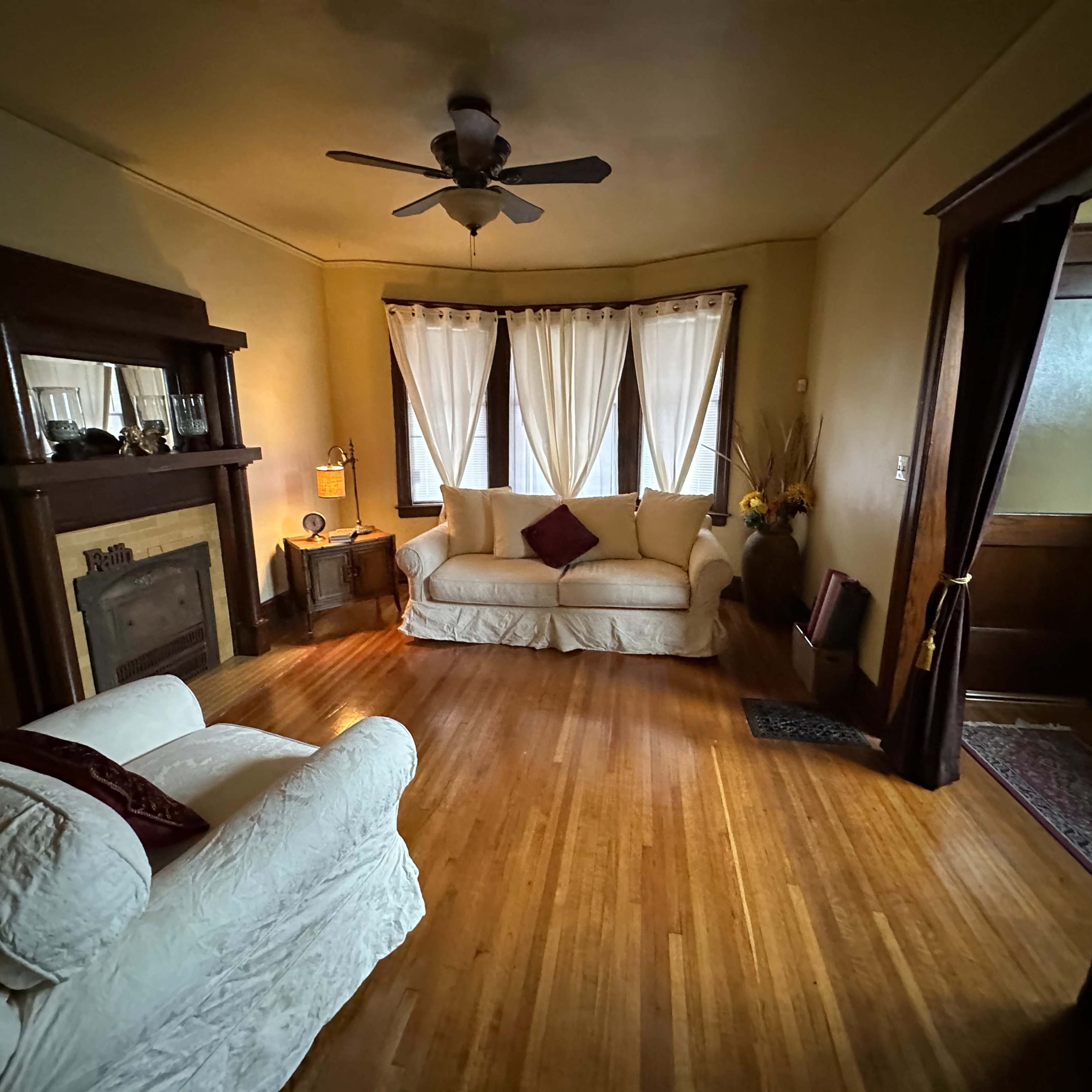 The room features two sofas, a fireplace, and large windows with sheer curtains, all set on polished wooden floors.