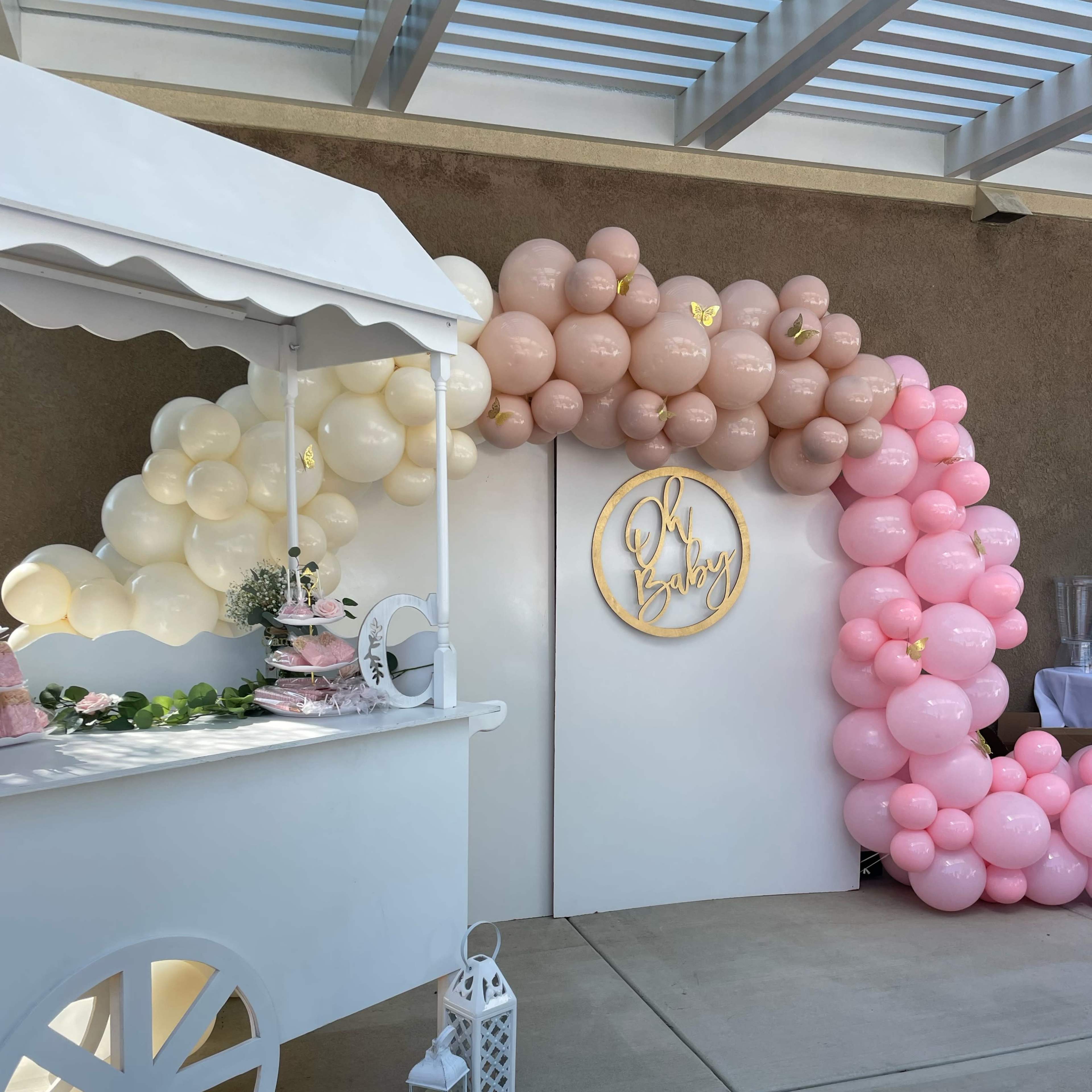 A decorated entrance with a white backdrop and a balloon arch in shades of pink and beige is complemented by a candy cart nearby.