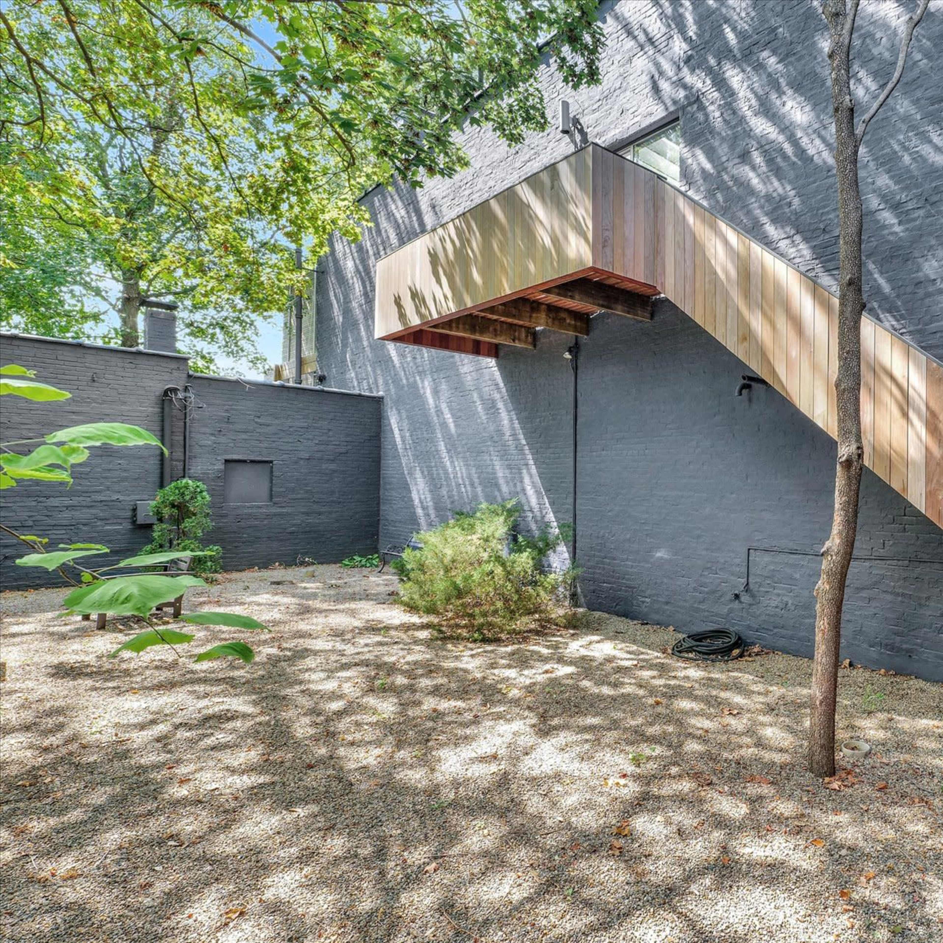 A wooden stairway leading to an upper entrance, beside a stone wall in a garden area with gravel and sparse greenery.