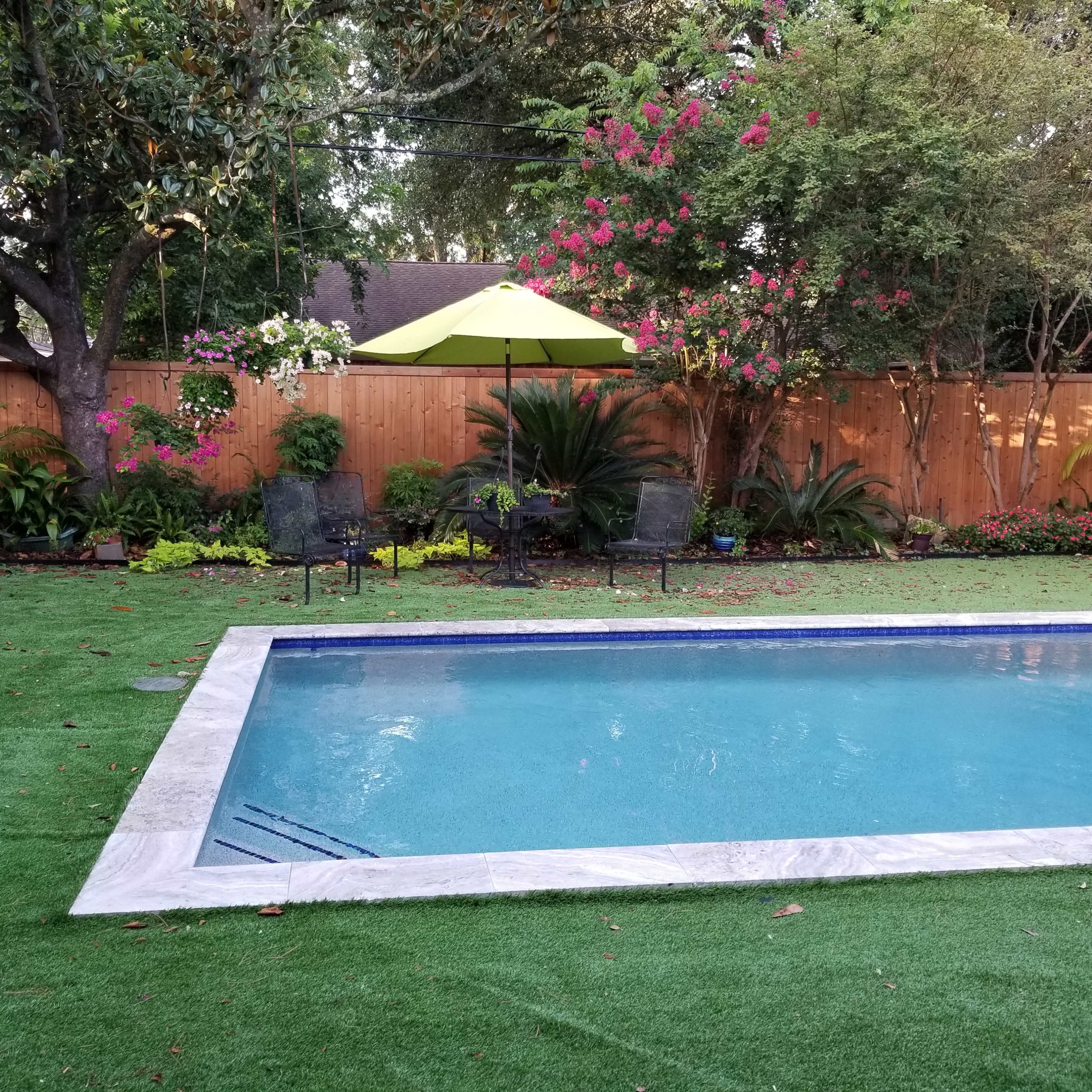 A landscaped backyard featuring a swimming pool surrounded by greenery, flowers, and outdoor seating.