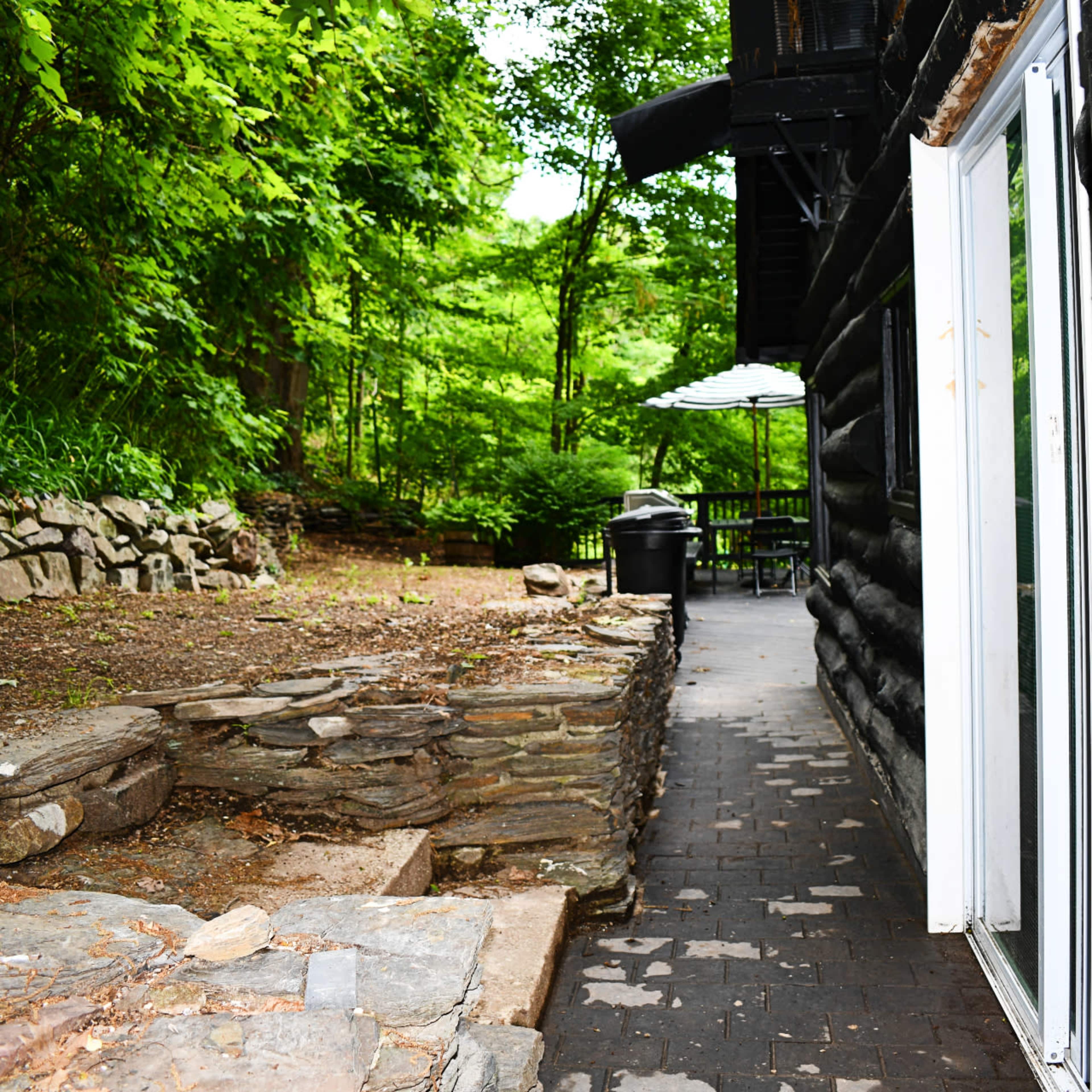 A stone pathway leads alongside a black log cabin, bordered by trees and a stone wall.