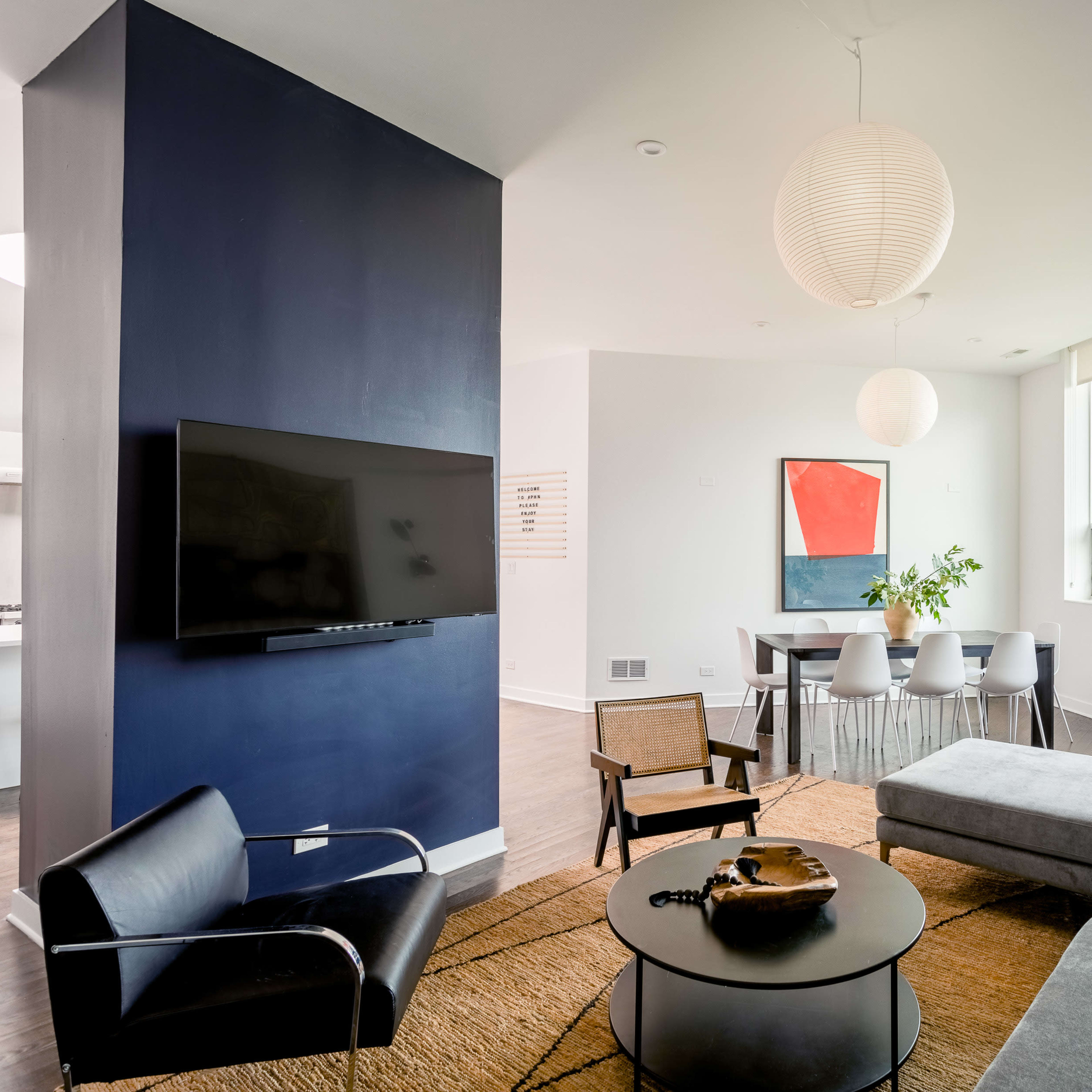A modern living space featuring a sectional sofa, a round coffee table, a wall-mounted television, and a dining area with a table and chairs, complemented by a large window and minimalist decor.