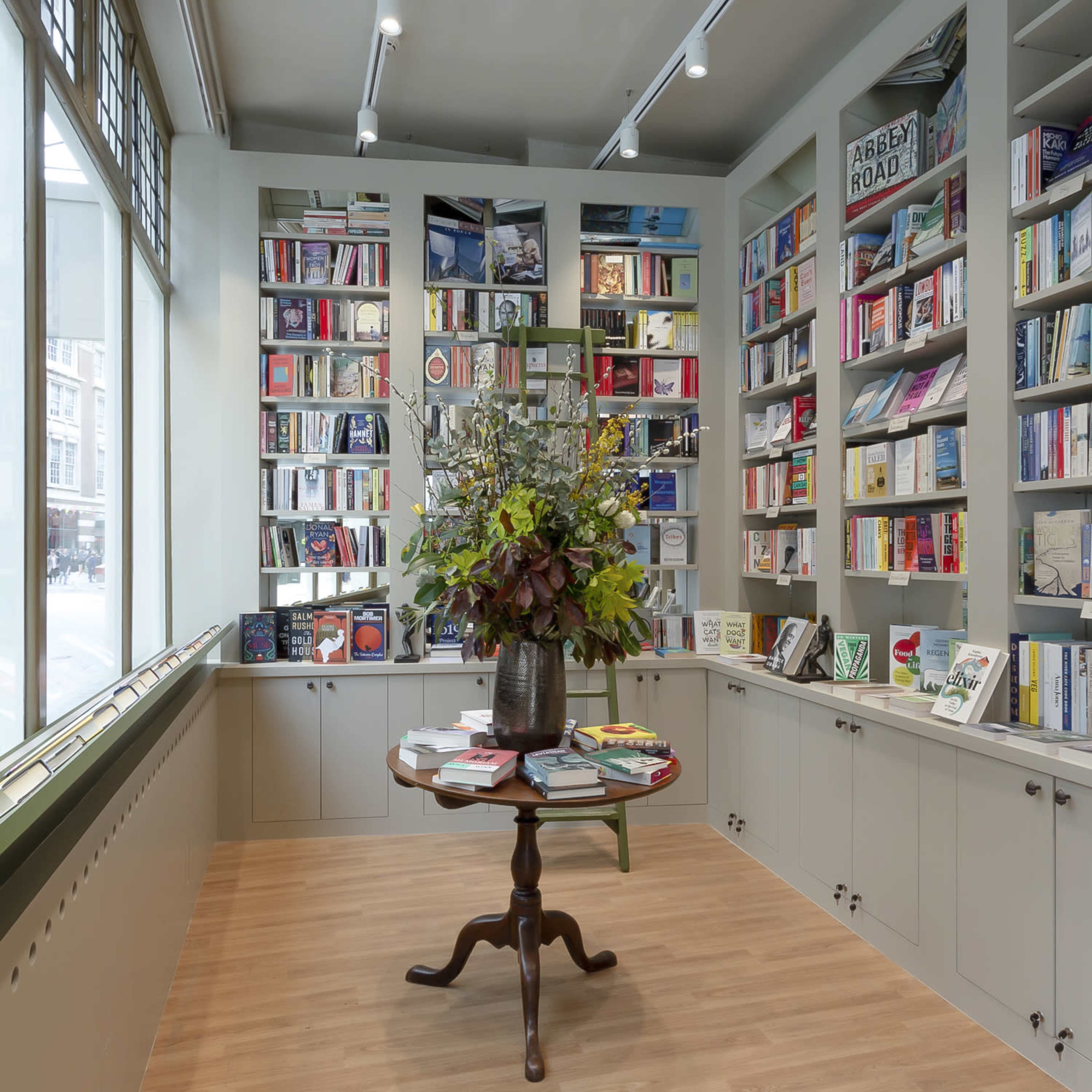 A cozy bookstore interior with shelves filled with books, a central round table adorned with a floral arrangement, and large windows allowing natural light to enter.