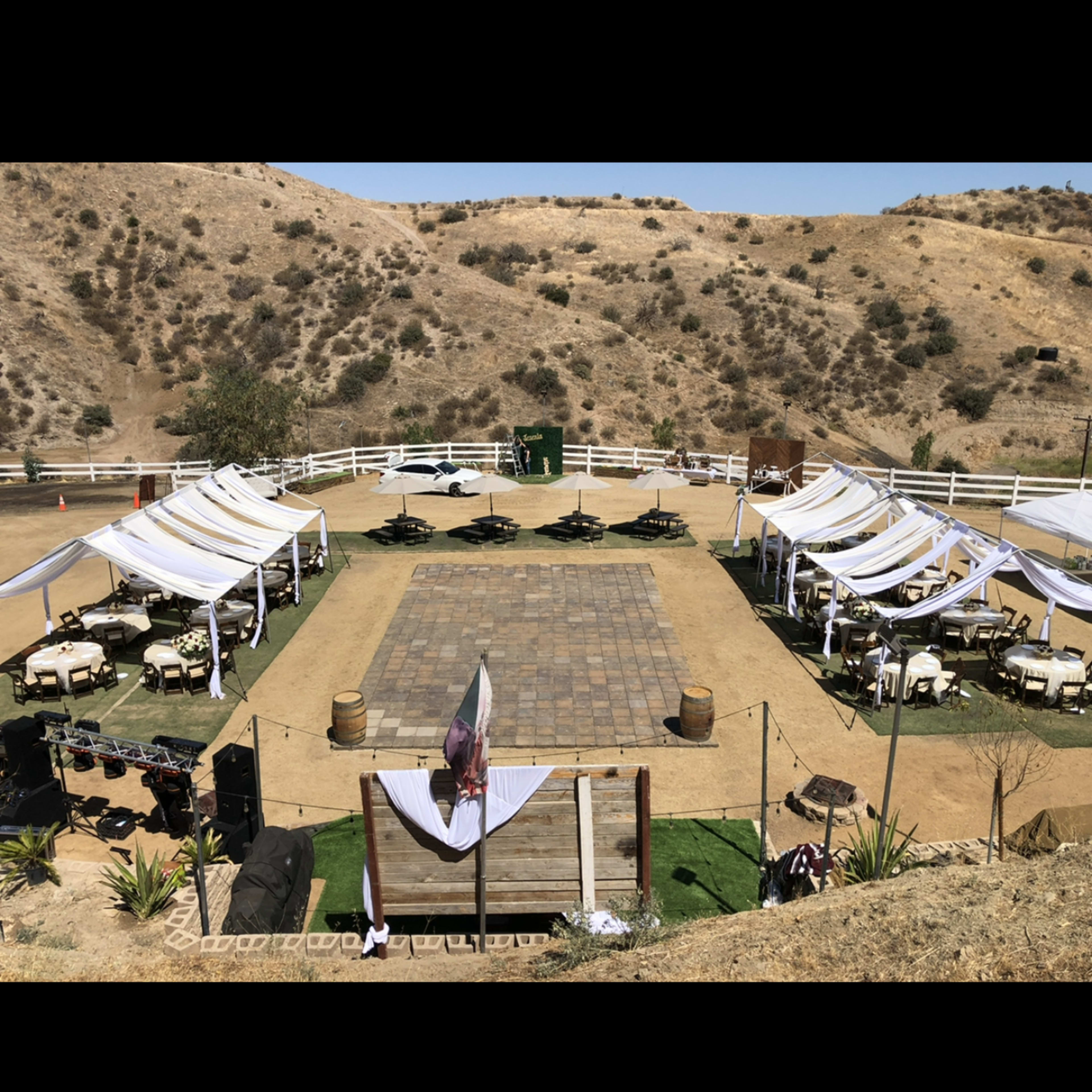 An outdoor event space set up with tables and tents arranged around a central dance floor in a hilly landscape.
