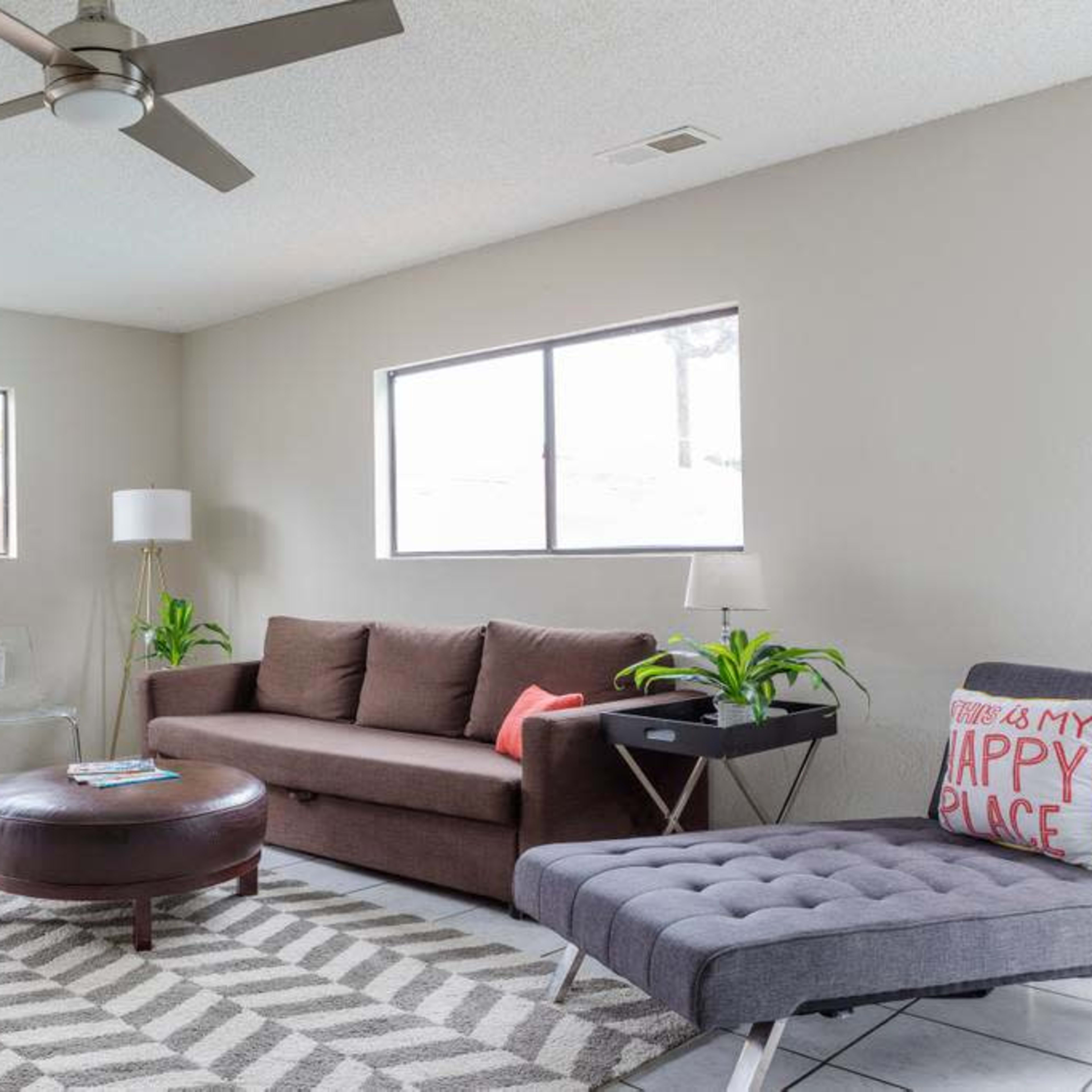 A living room features a brown sofa, a gray chaise lounge, a glass side table, and plants, with two windows allowing natural light.