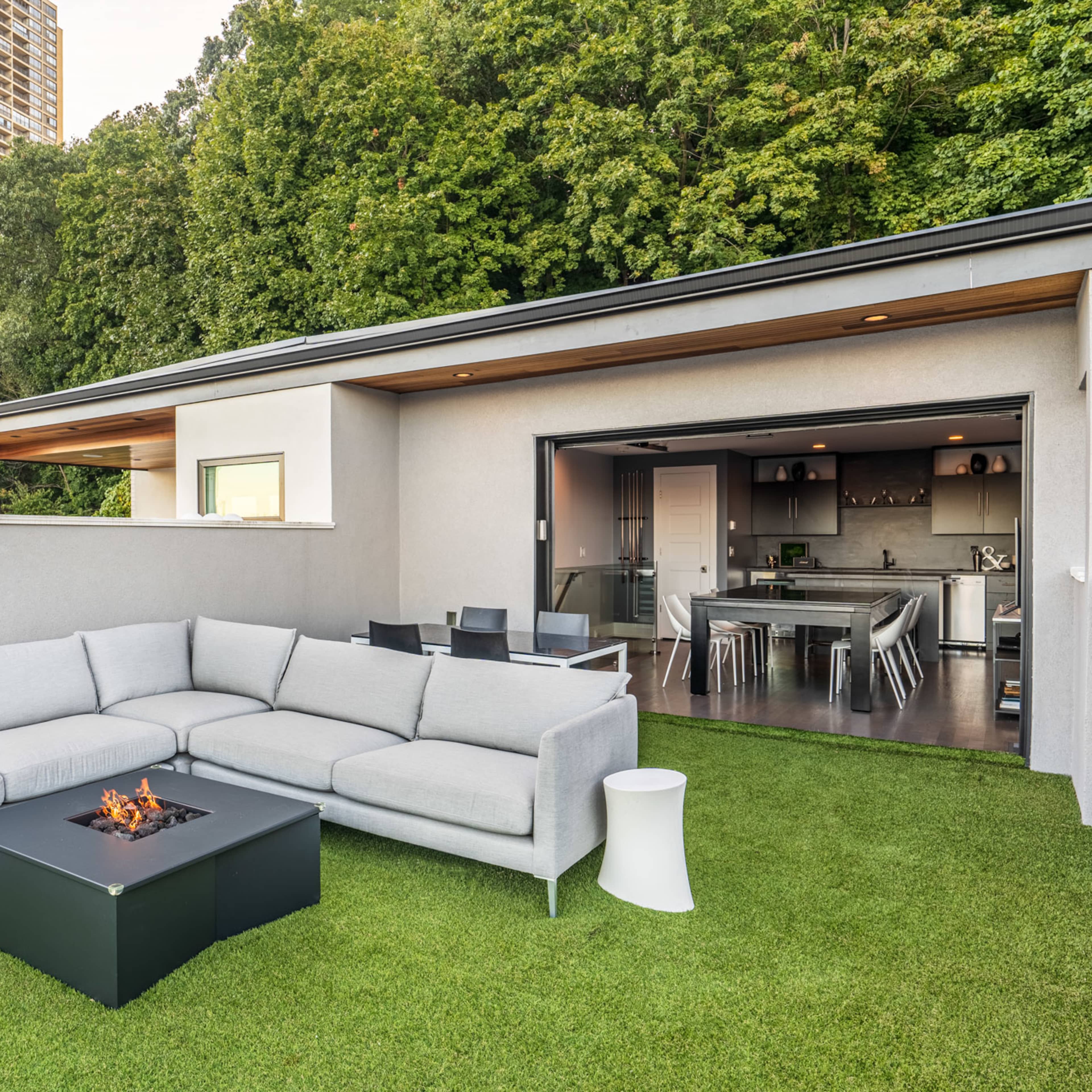 A modern outdoor seating area with a gray sectional sofa and a fire table on an artificial lawn, adjacent to an open-concept indoor space.