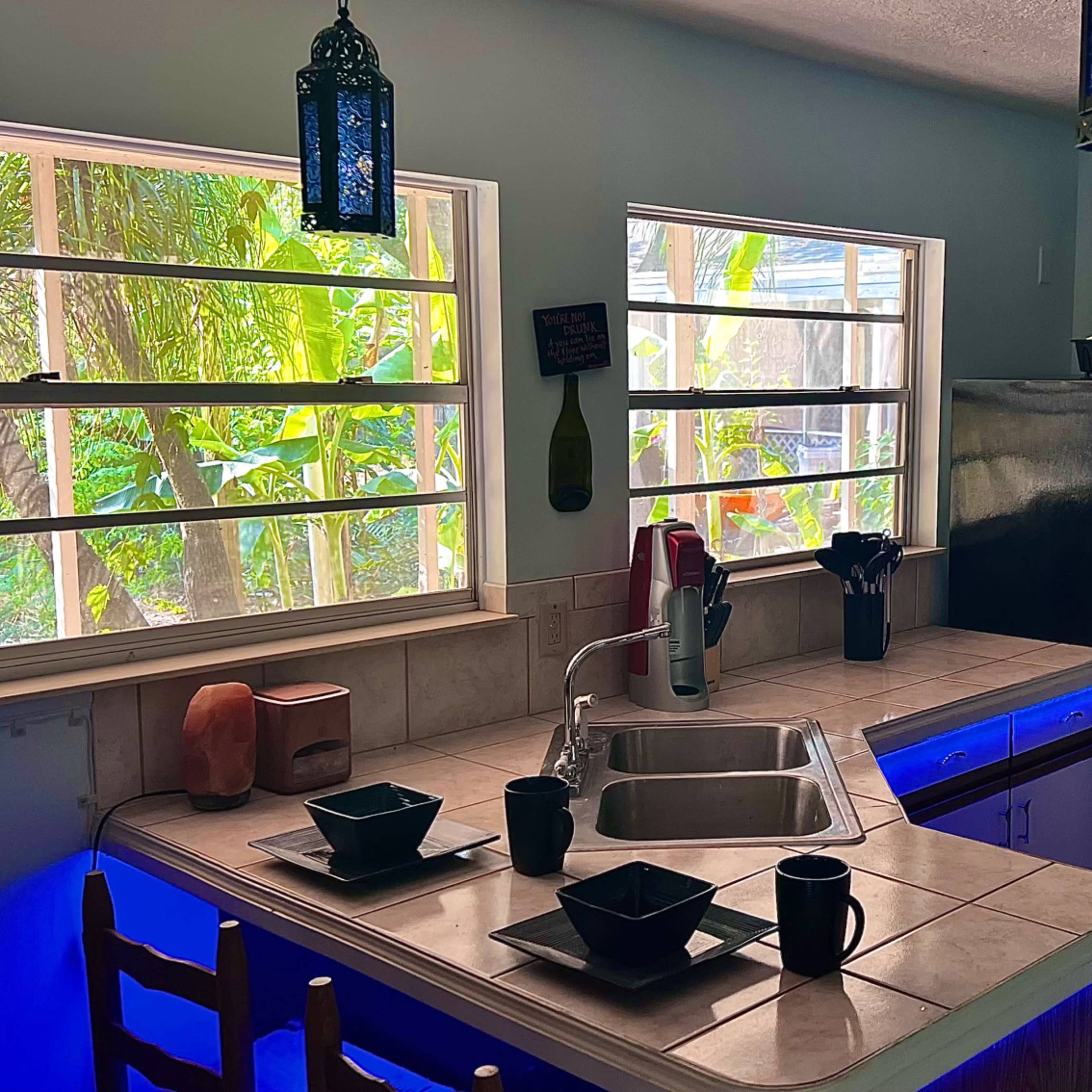A kitchen with a tiled countertop, stainless steel refrigerator, and large windows that overlook green foliage outside.