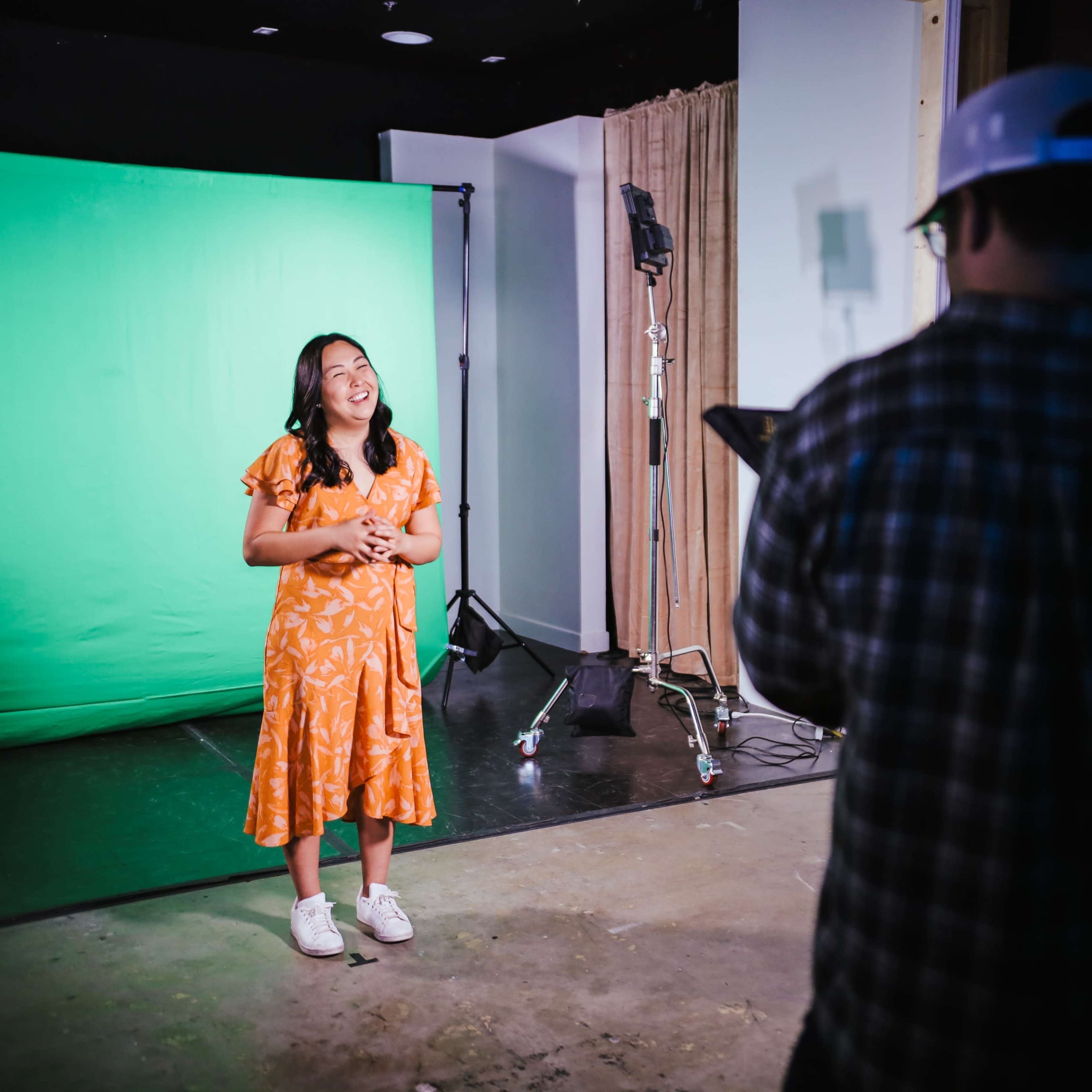 A person in an orange dress stands in front of a green screen while another person, wearing a plaid shirt and a cap, observes nearby.