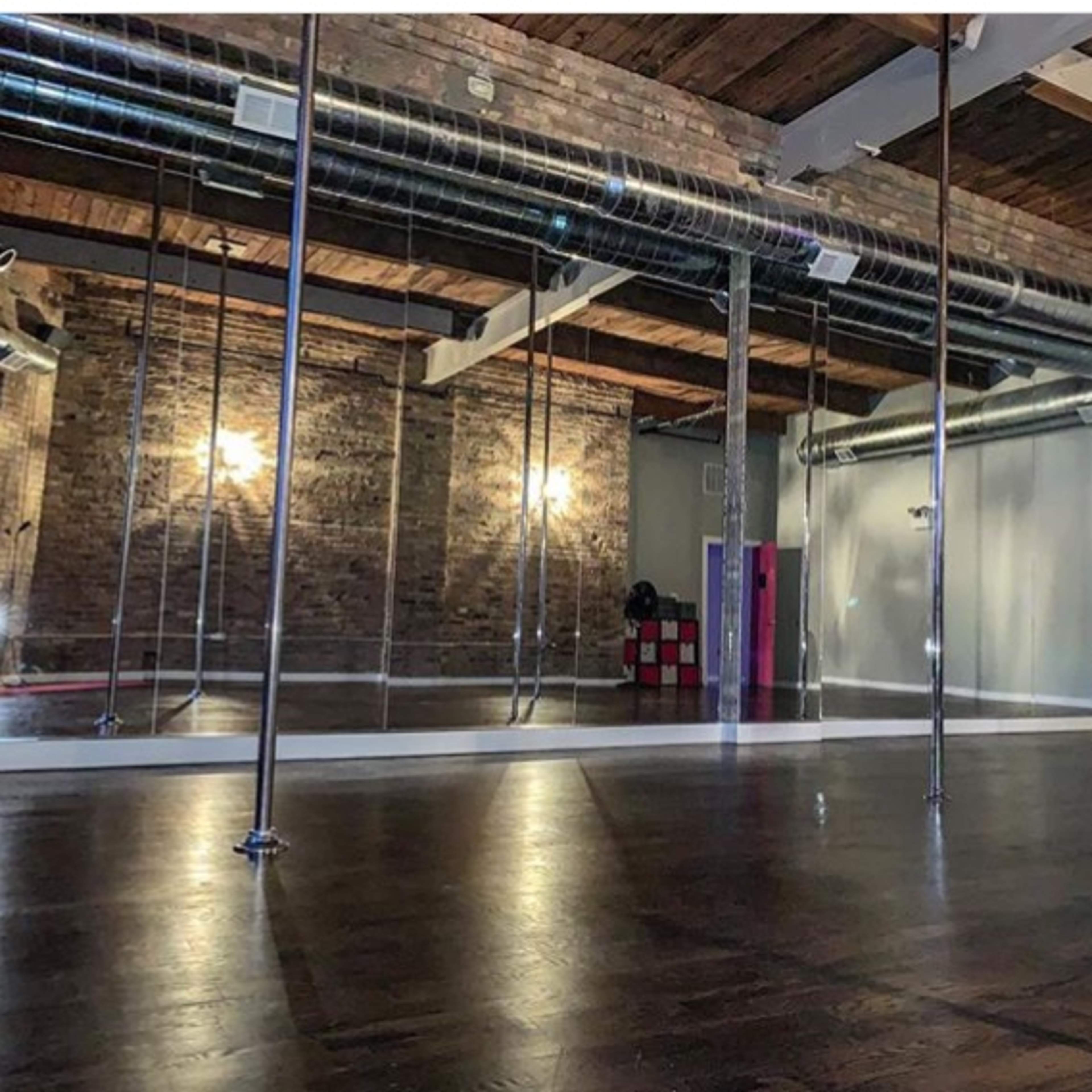 An empty dance studio featuring polished wooden floors, mirrored walls, and several pole dance installations.