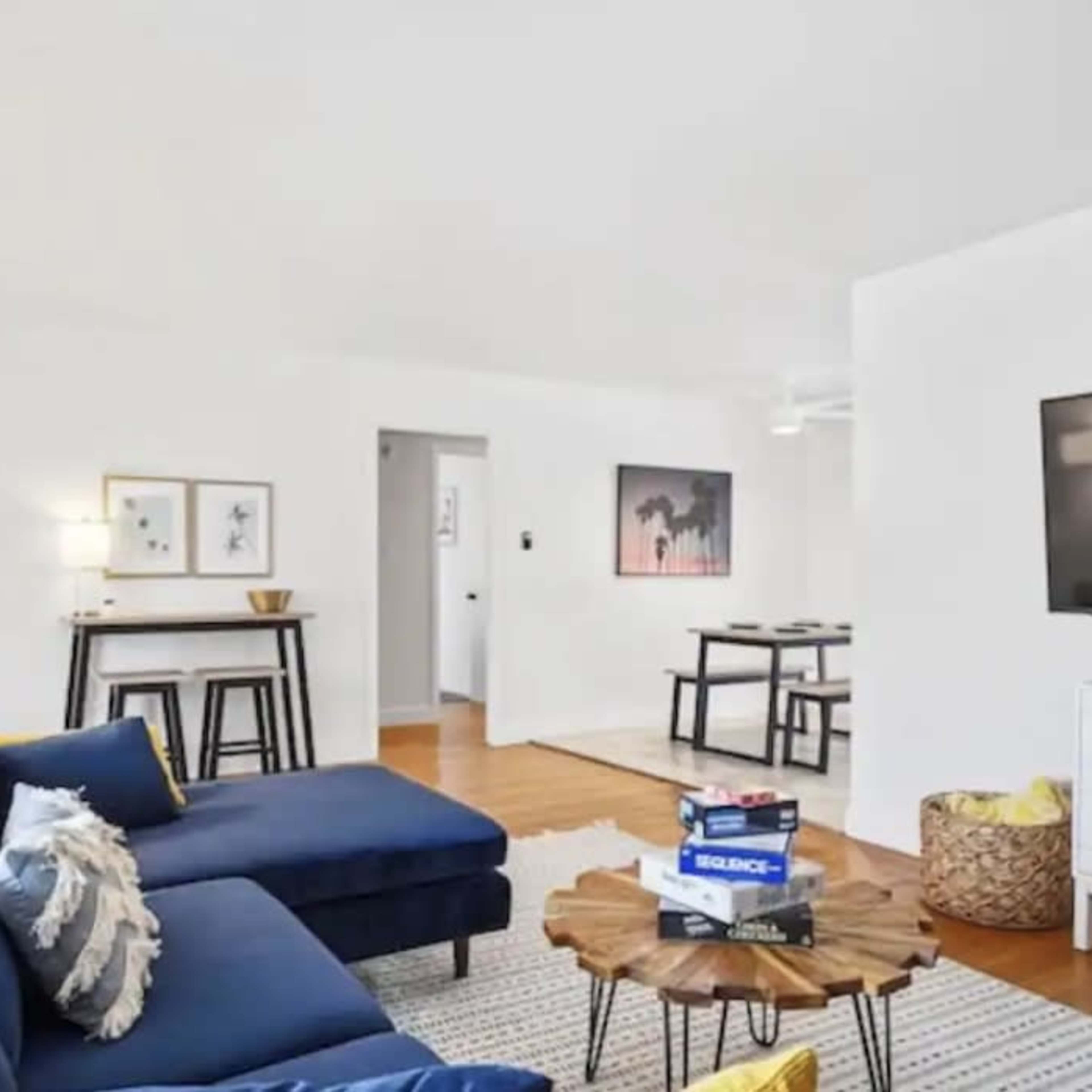 A brightly lit living room with a blue sectional sofa, a wooden coffee table, a television on a white cabinet, and a dining area in the background.