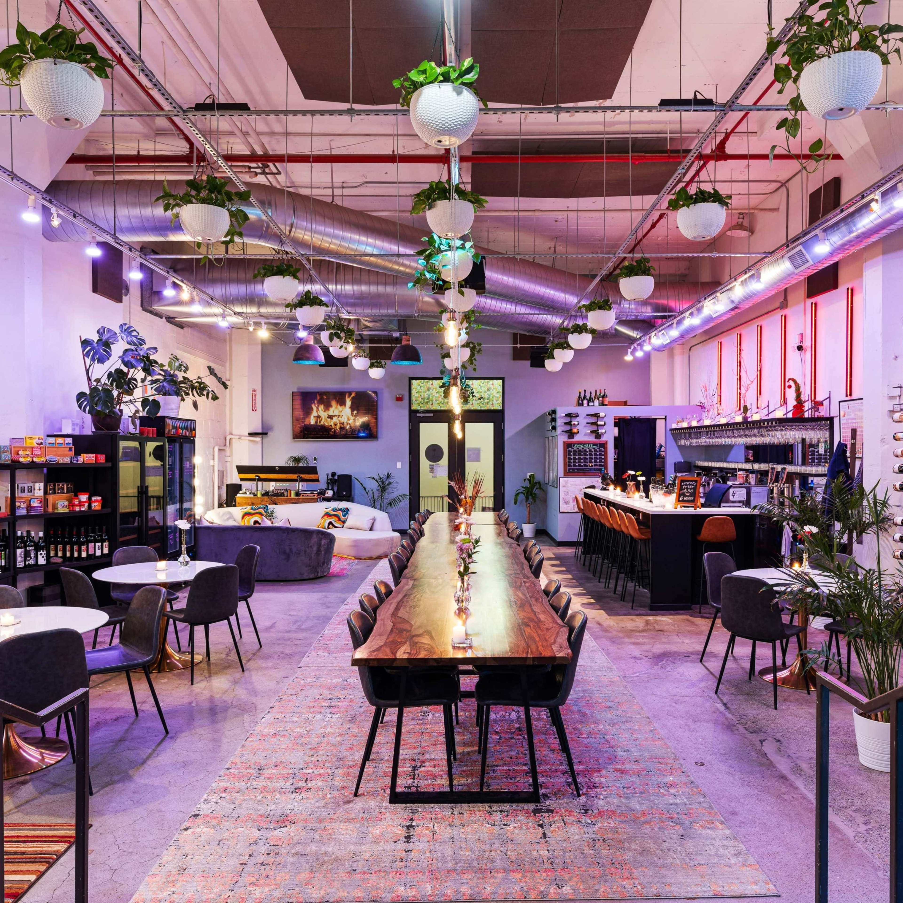 A modern, spacious restaurant with a long wooden dining table, surrounded by plants, shelves of wine, and various seating arrangements.