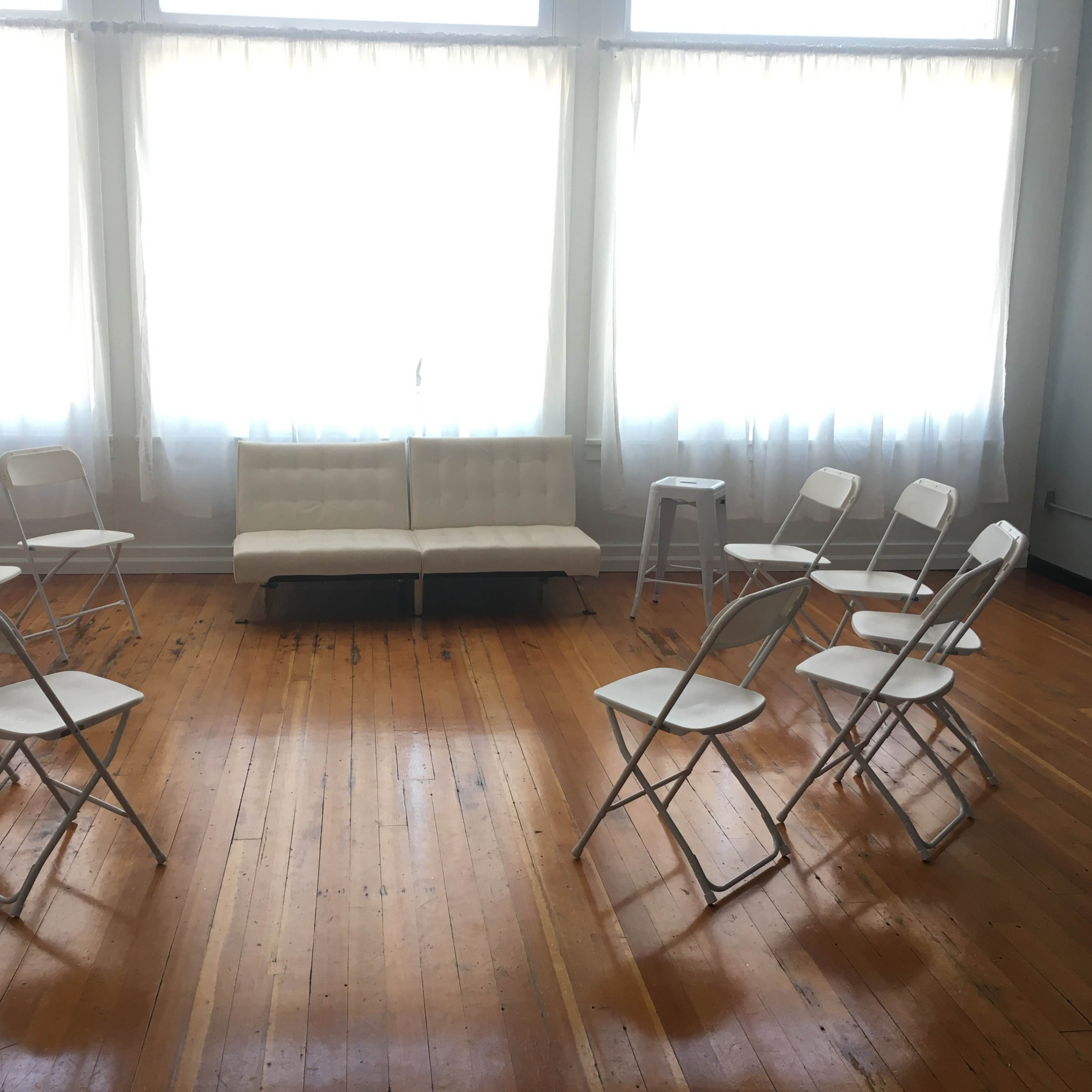 A room with large windows and natural light, featuring a circular arrangement of white folding chairs around a white couch and a small stool.