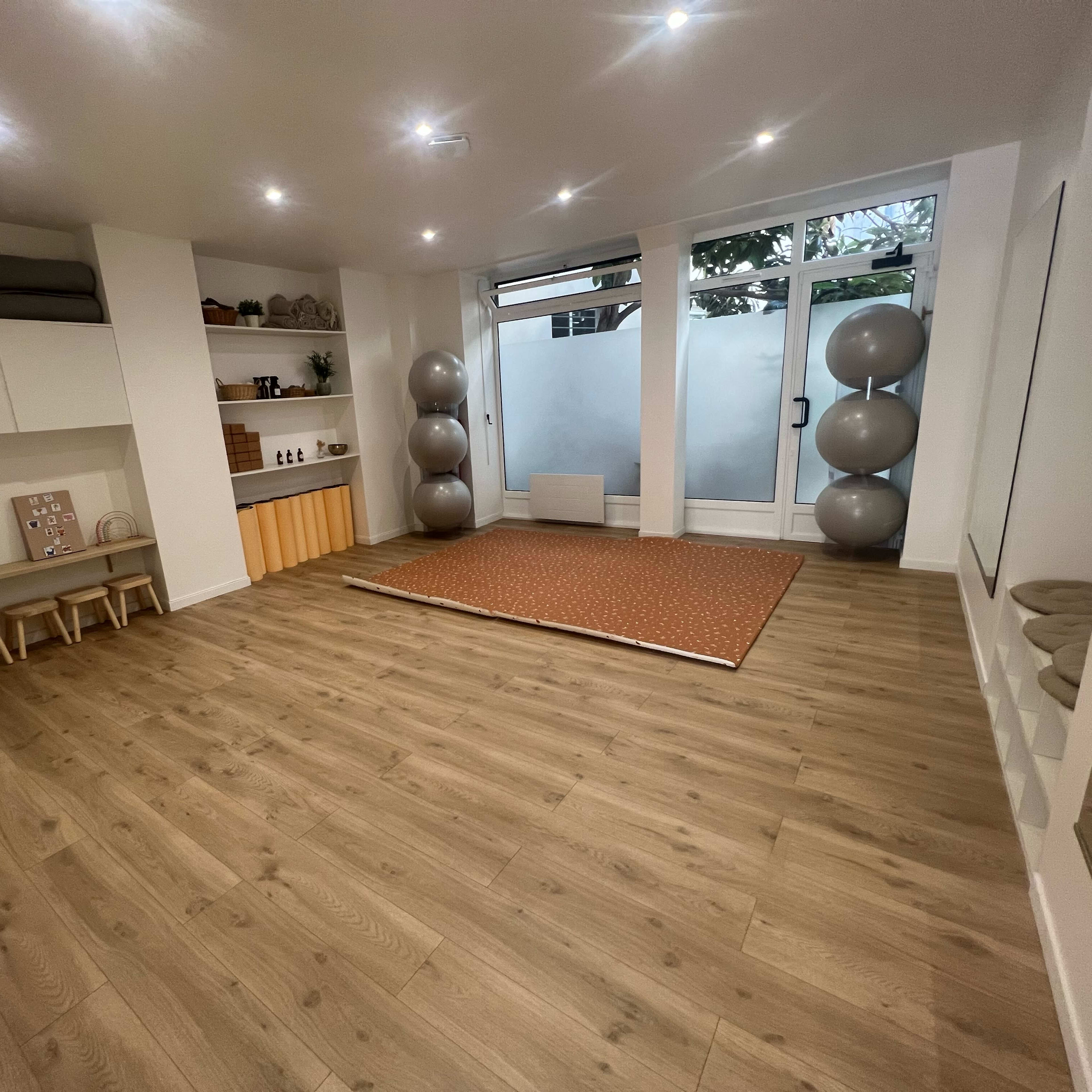 A spacious, well-lit room with wooden flooring, minimal furniture, and large windows.