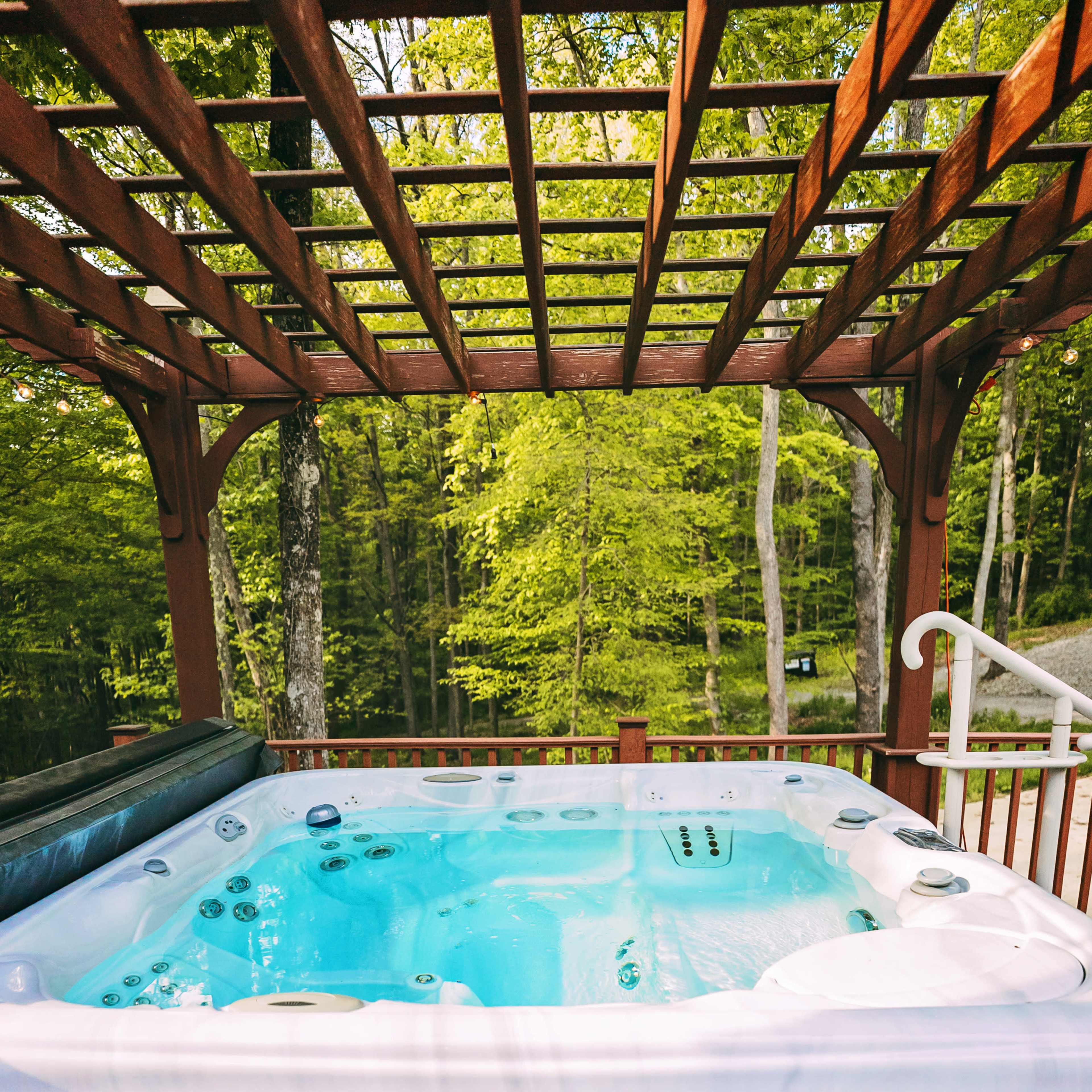 A white hot tub with a wooden deck is located under a pergola surrounded by lush green trees.