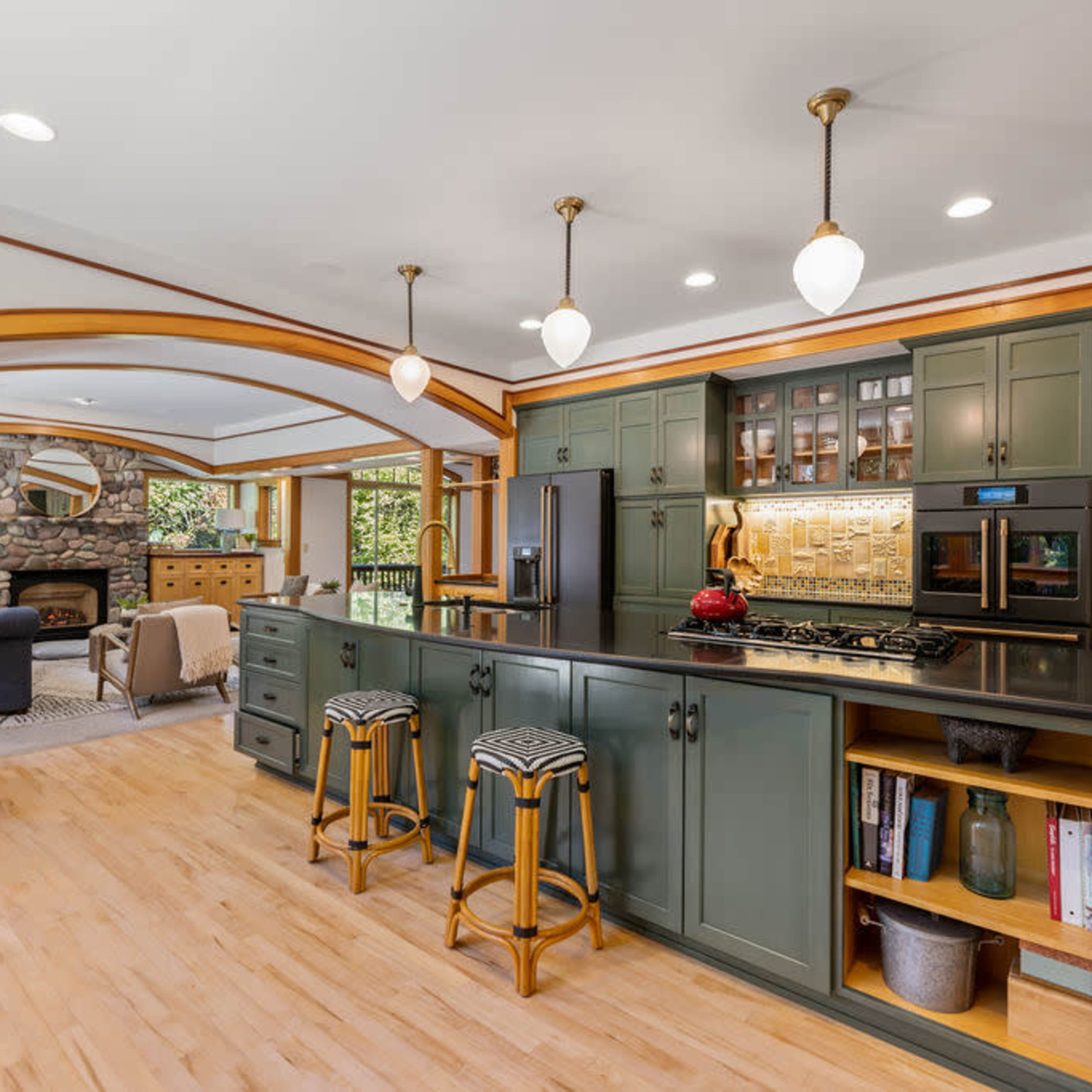 A modern kitchen with green cabinetry, a black countertop, and pendant lighting, adjacent to a living area with large windows and a stone fireplace.