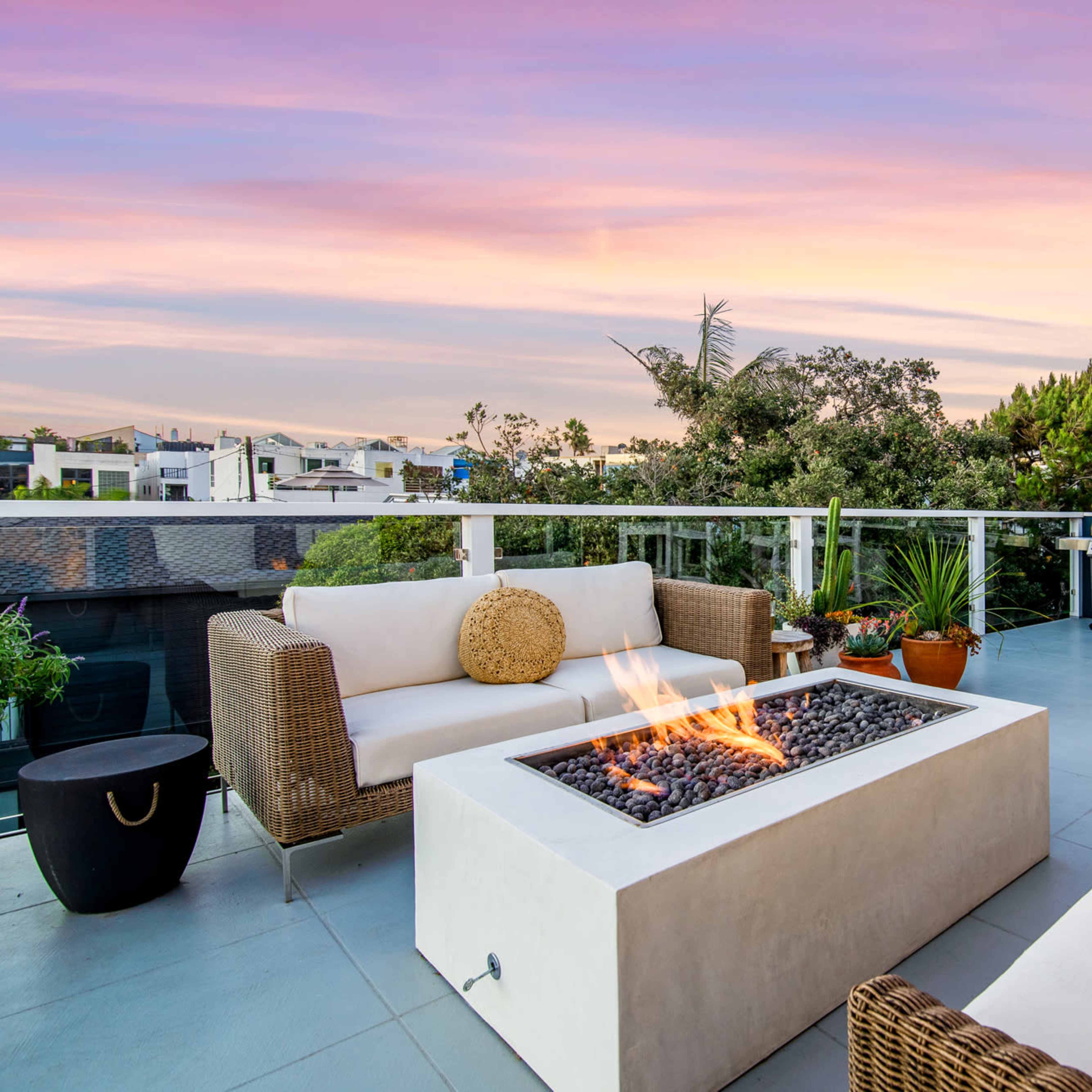 A modern outdoor patio features two wicker sofas surrounding a fire pit, with a dining area and vibrant sunset in the background.
