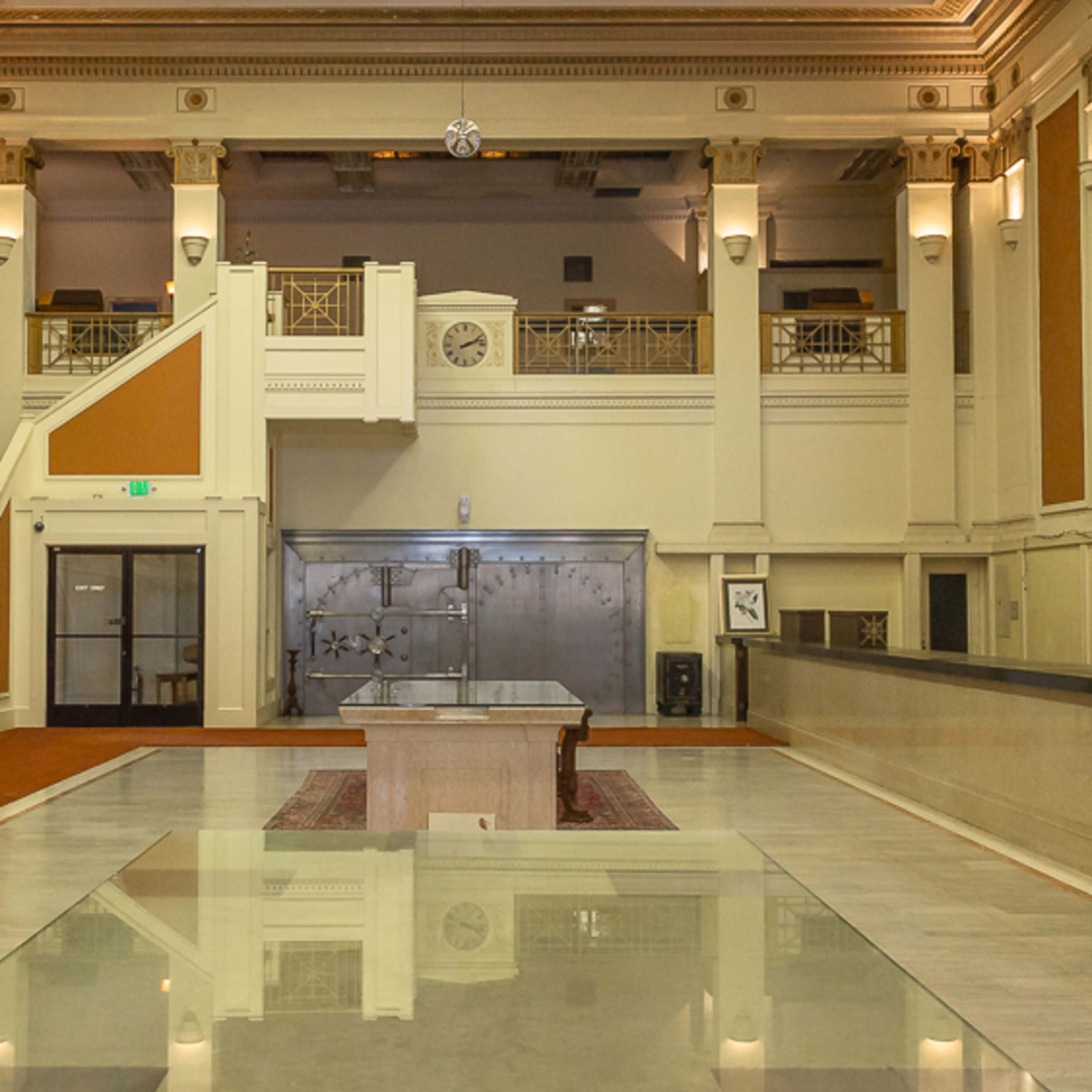 An empty grand hall with high ceilings, a stairway on the left, and a large vault door in the background.