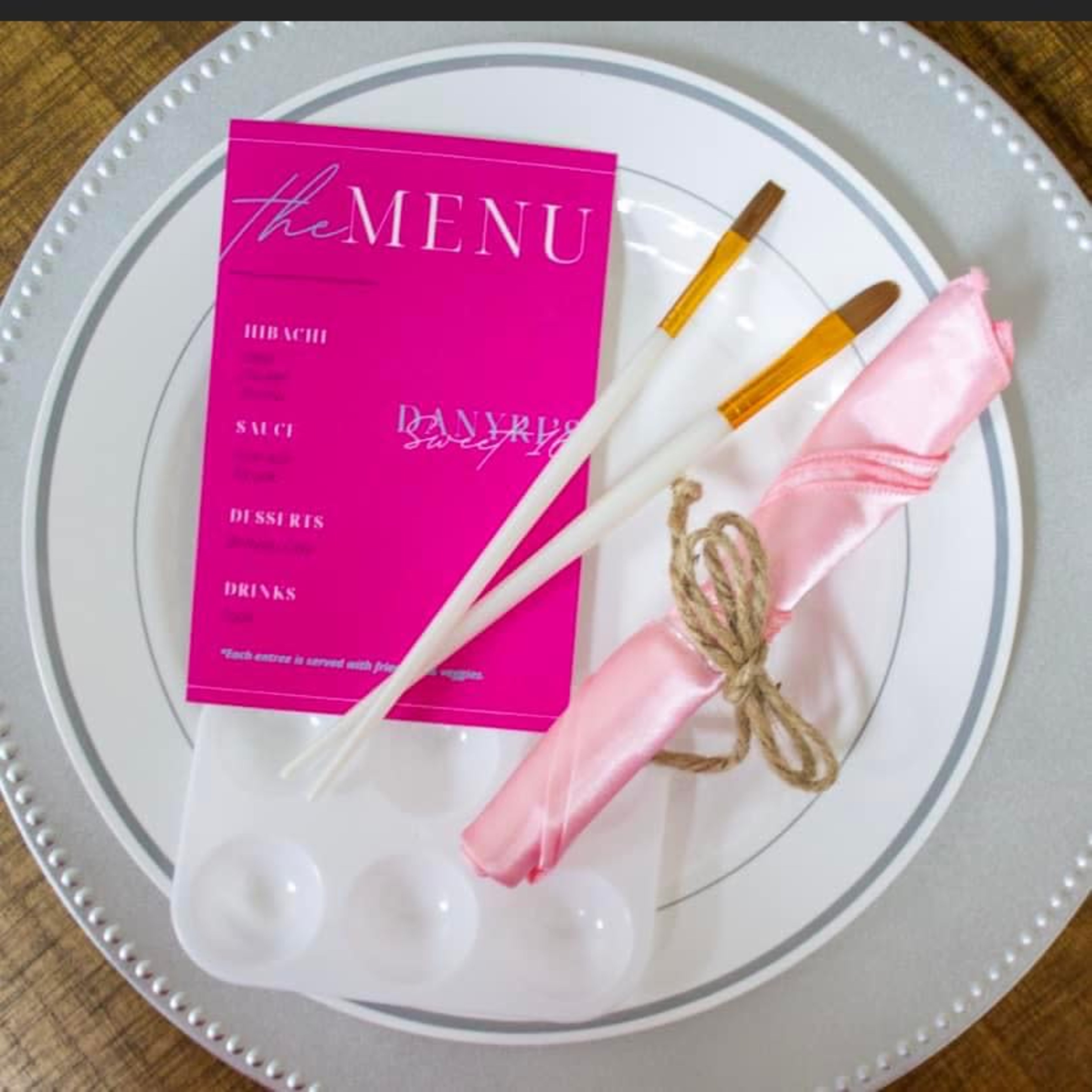 A set table features a pink menu card, decorative chopsticks, and a pink napkin tied with twine on a white plate.
