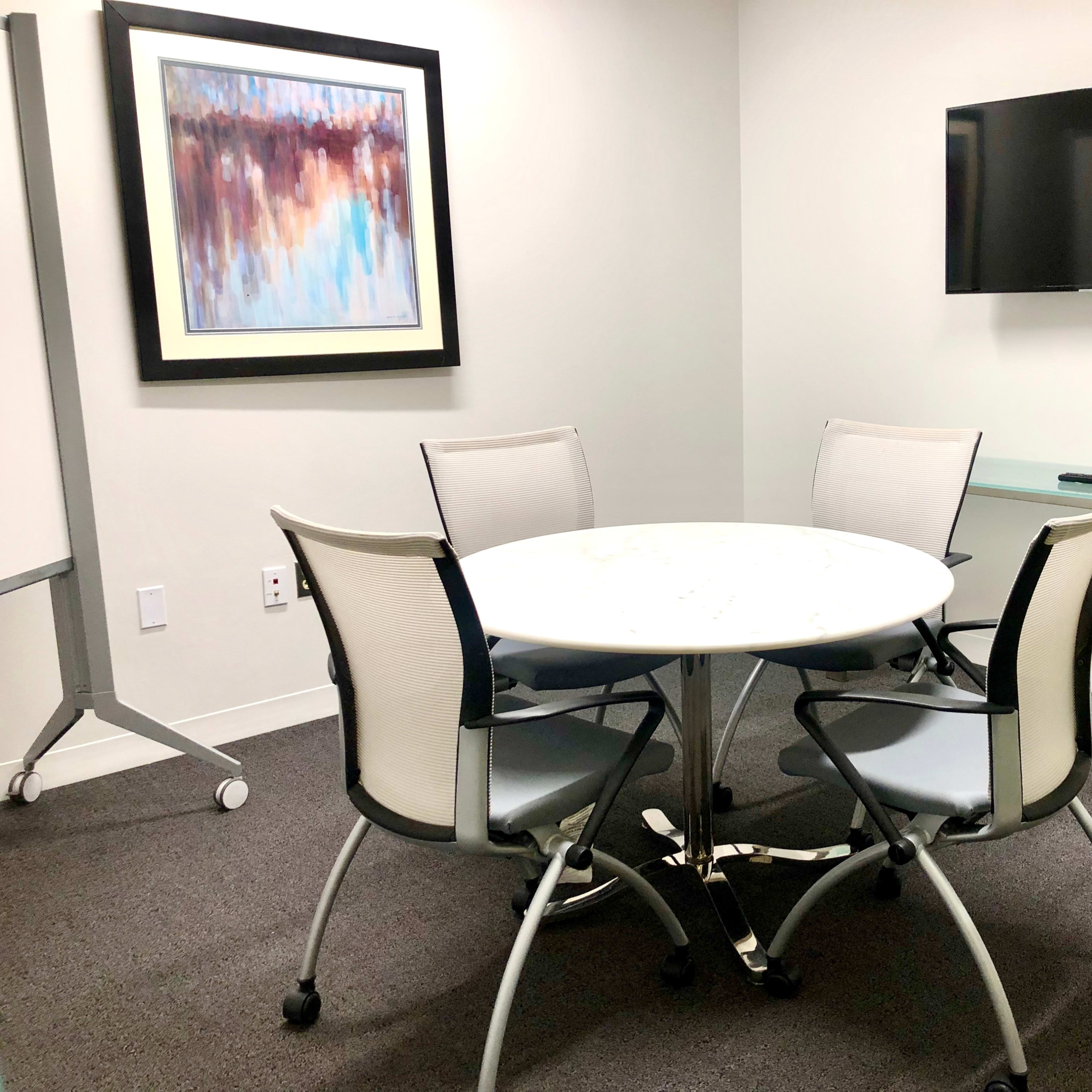 A small meeting room featuring a round table with four chairs, a flat-screen TV mounted on the wall, and a piece of abstract artwork.
