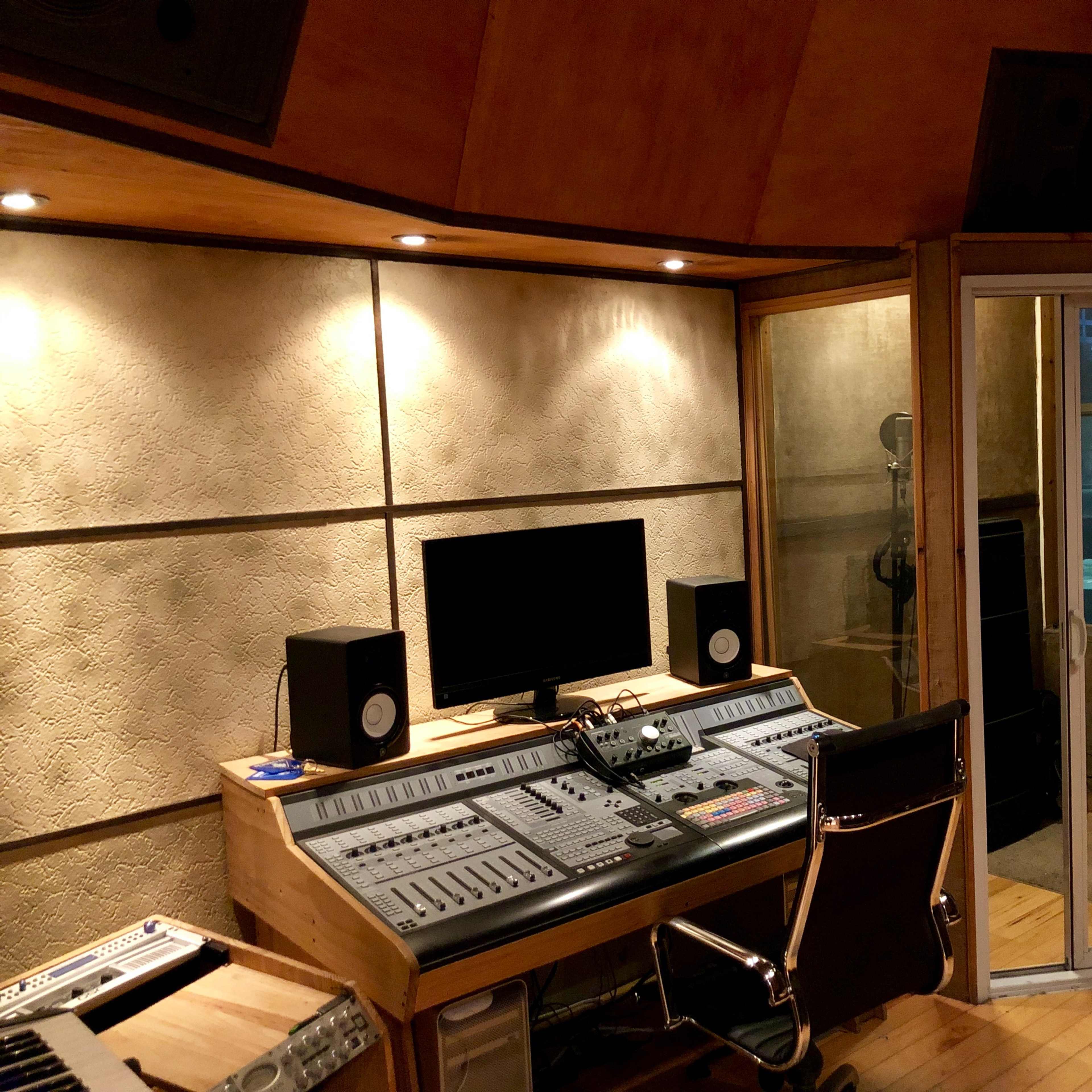 A music recording studio featuring a control desk with a computer monitor, speakers, and a glass-walled vocal booth.