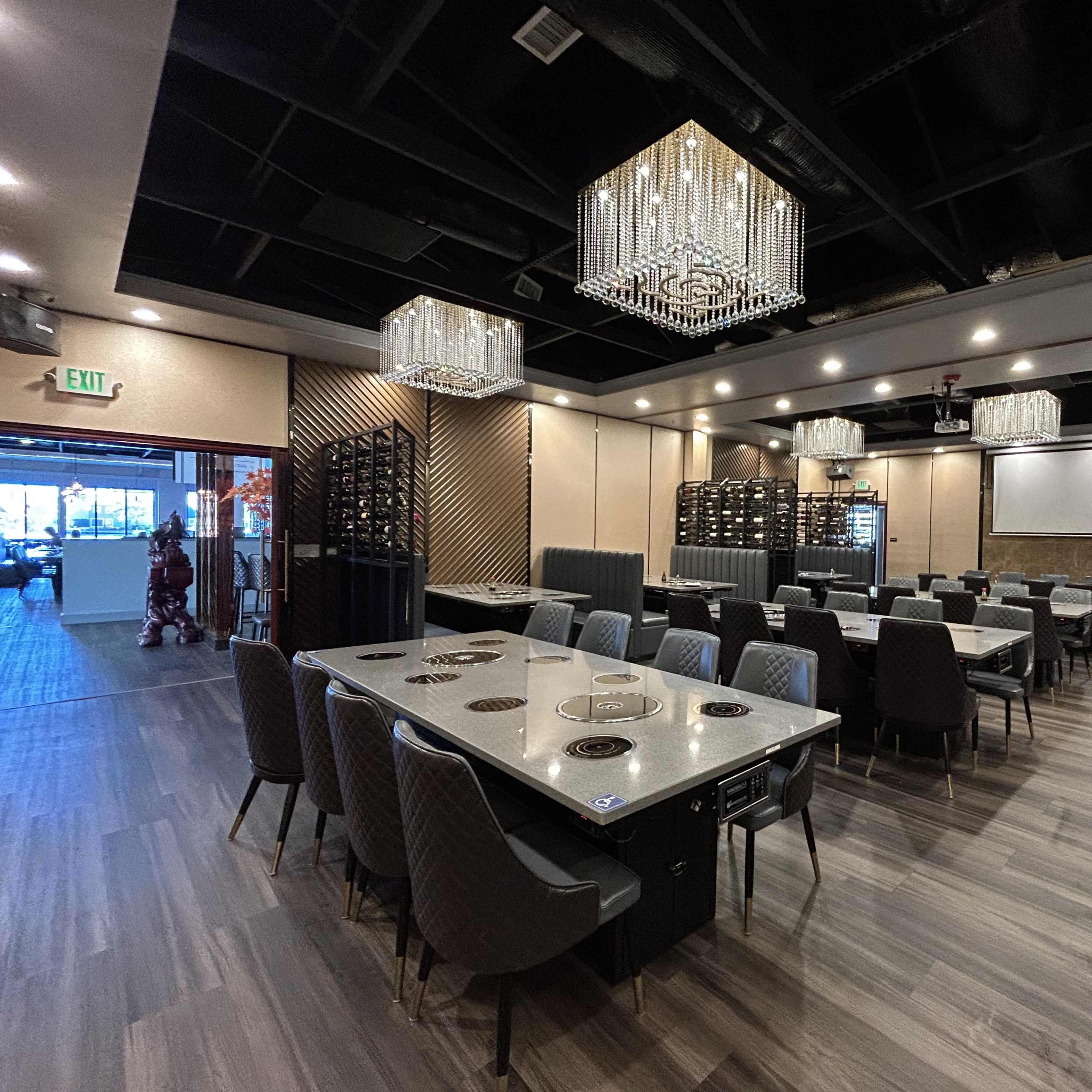 A modern restaurant interior featuring tables with built-in grilling stations, elegant chandeliers, and a wine rack in the background.