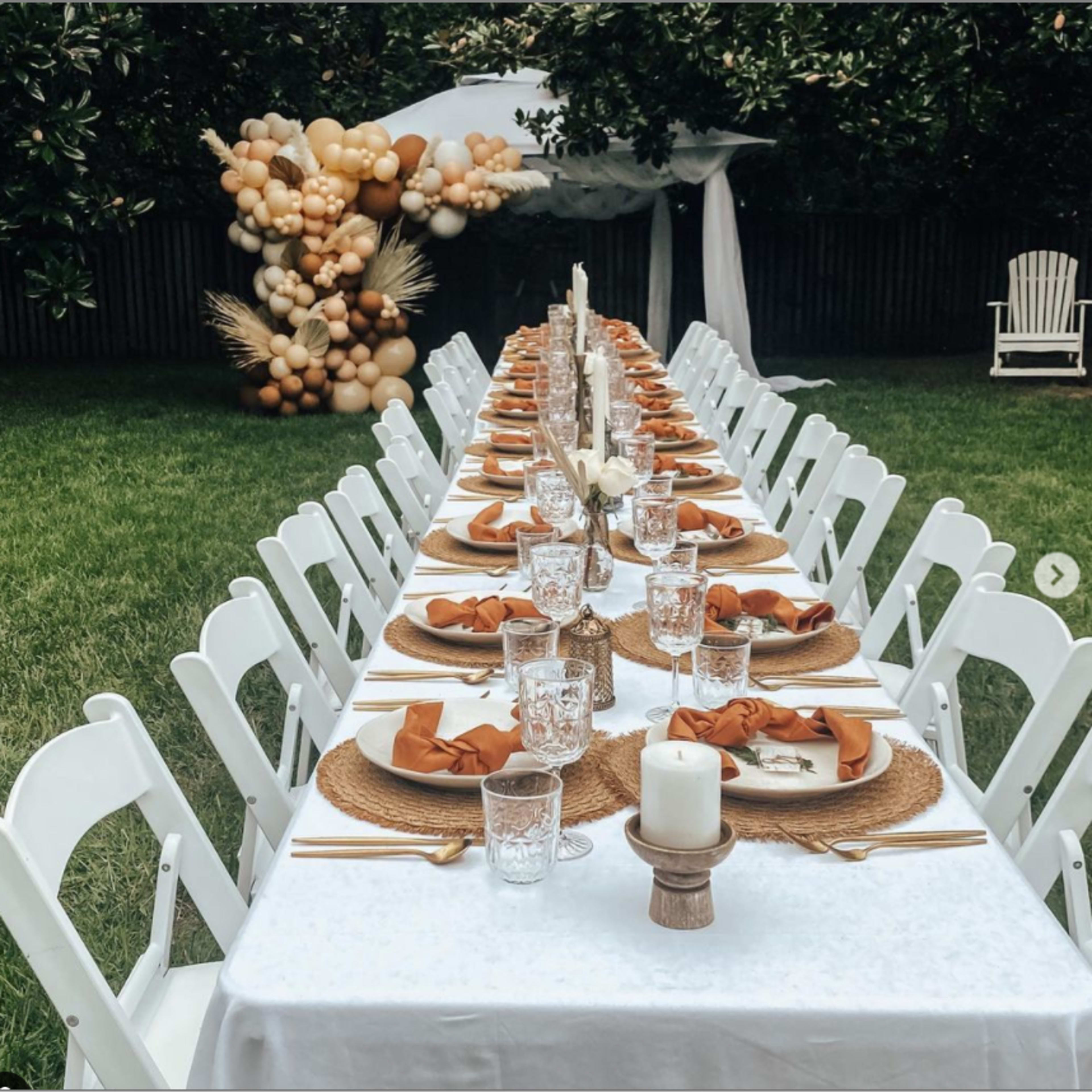 A long, elegantly set outdoor dining table is arranged on a green lawn, flanked by white chairs and decorated with neutral-toned tableware and a backdrop of a balloon arch.