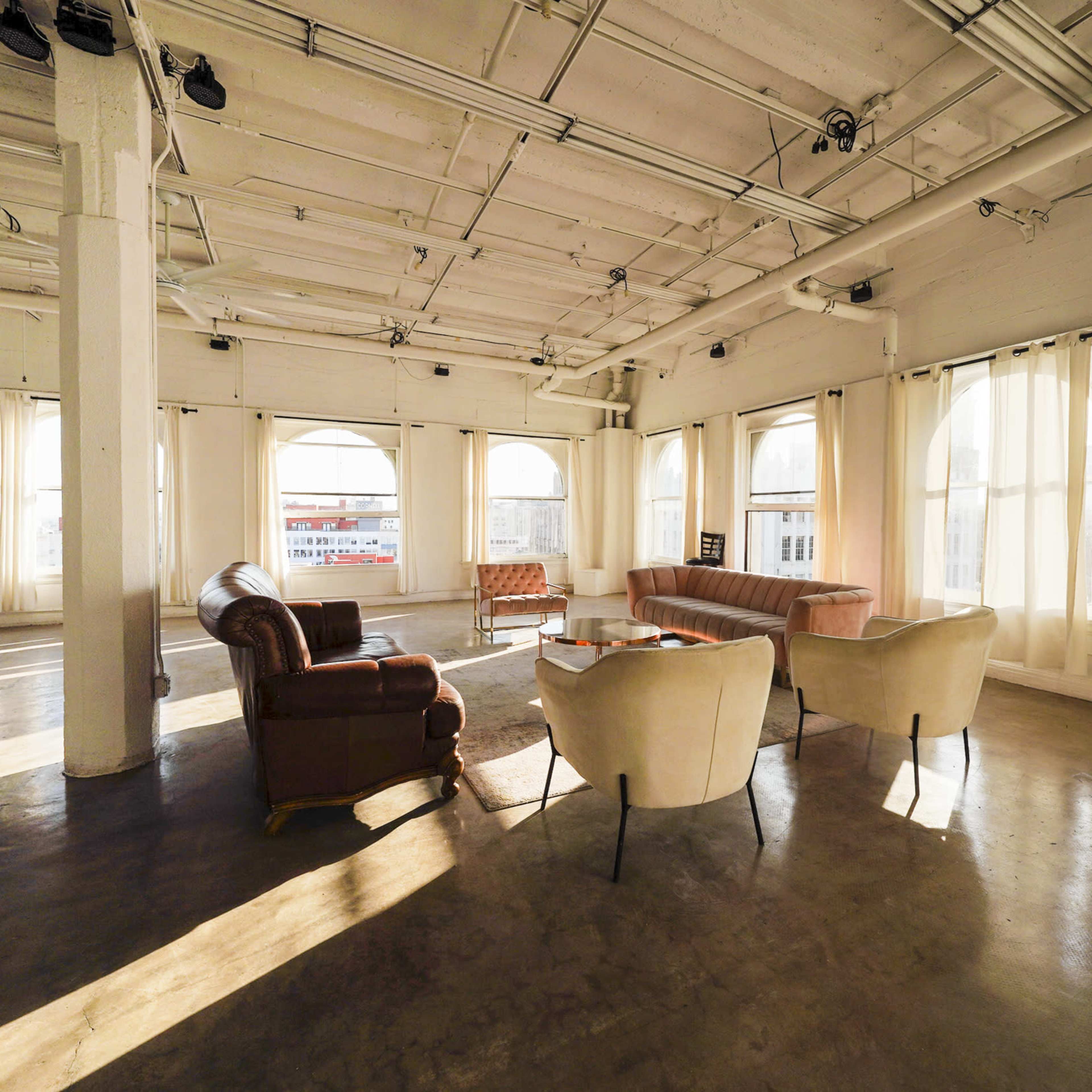 A spacious, well-lit room with large windows, featuring a mix of upholstered seating arrangements and a coffee table.