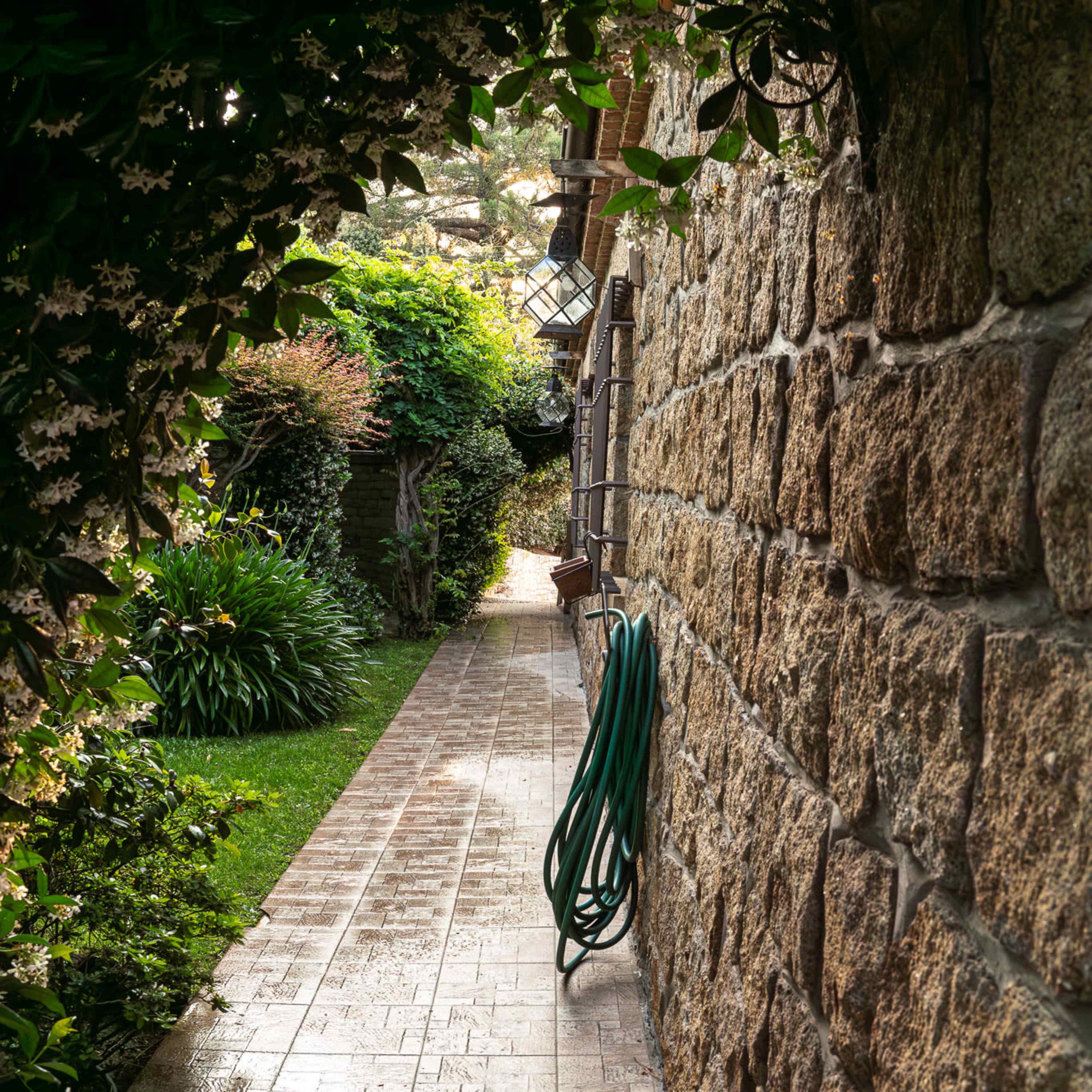 A narrow pathway bordered by lush greenery and flowering vines leads through a stone archway.