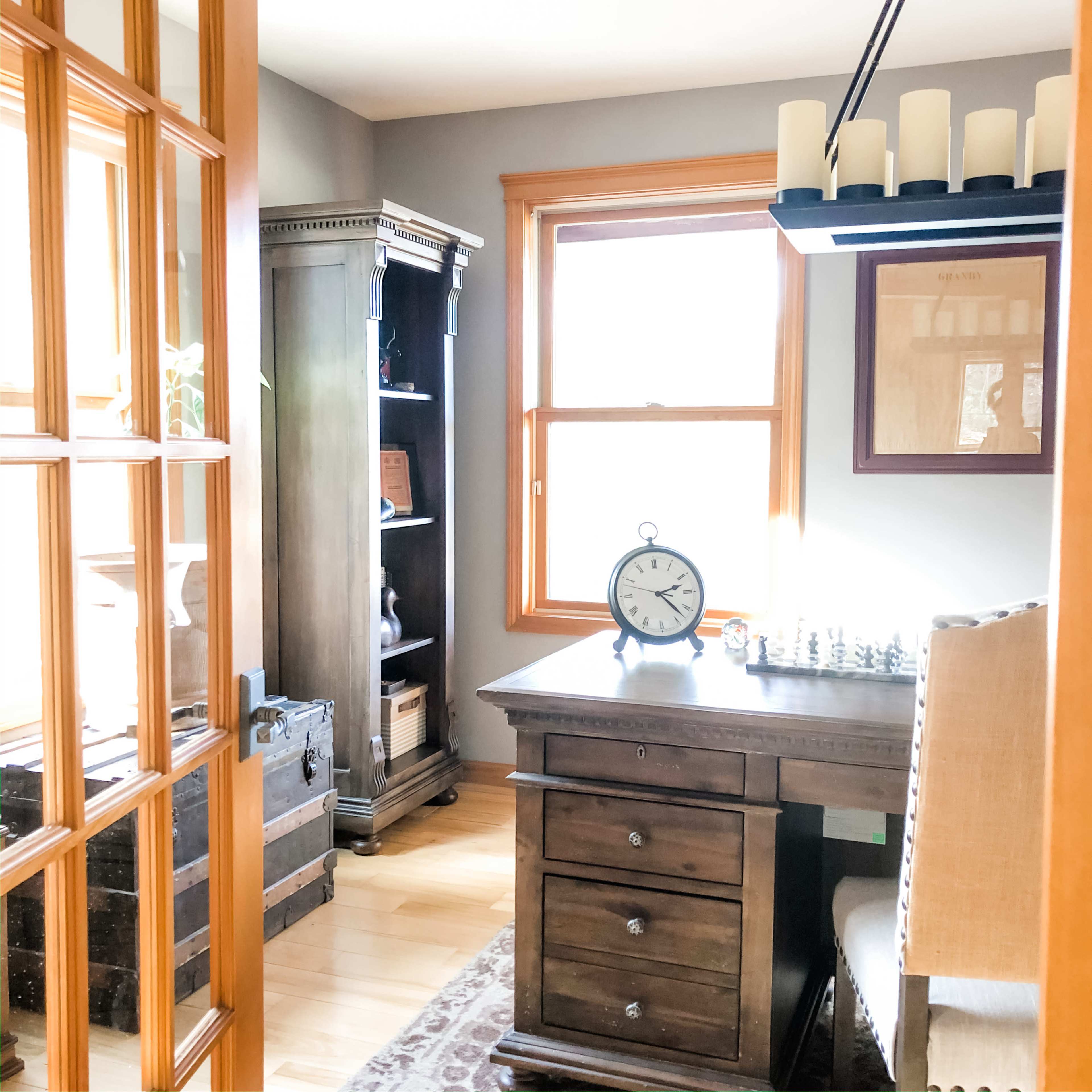 A well-lit office space featuring a wooden desk, a clock, a chessboard, and a bookshelf against a window.
