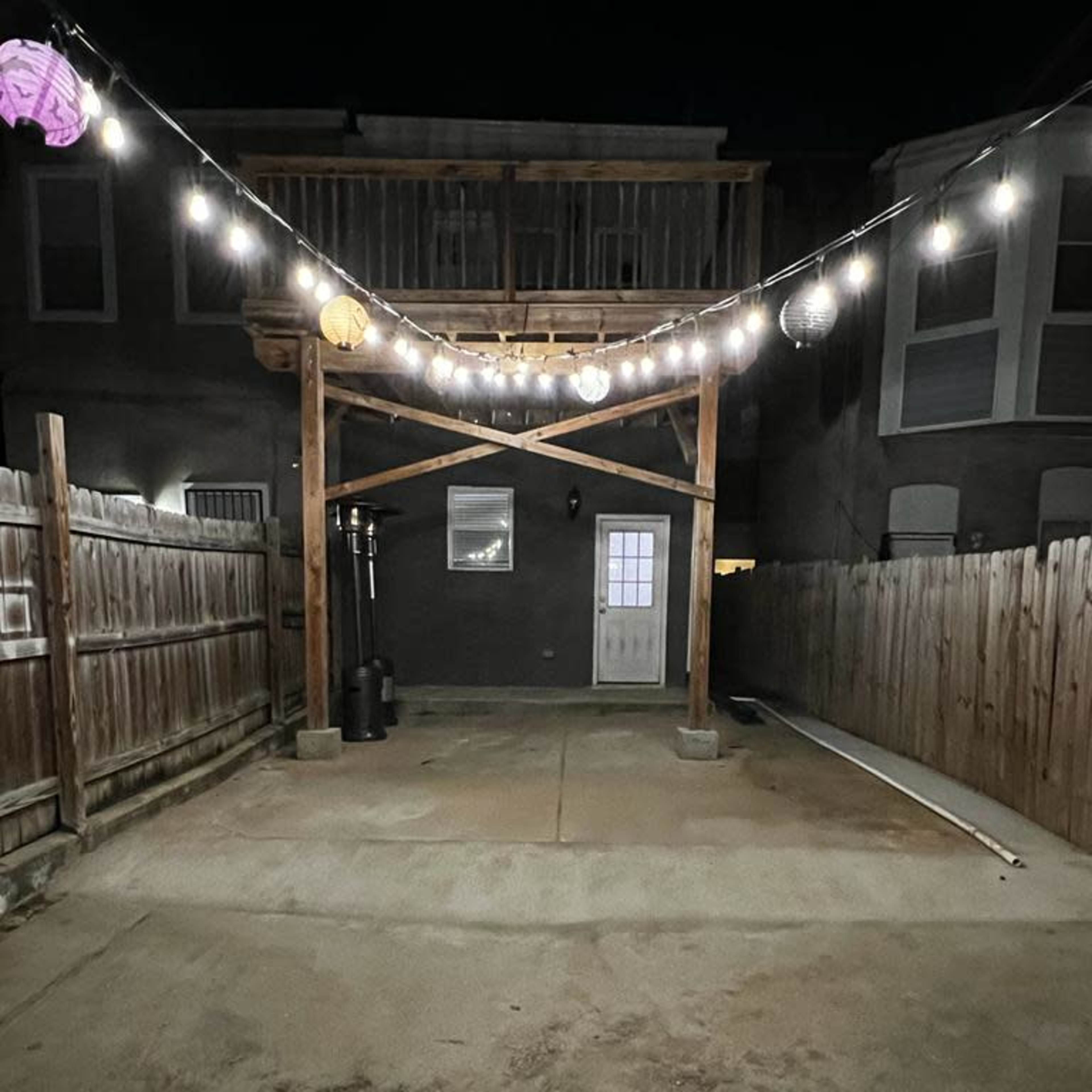 A dimly lit backyard area with a wooden structure overhead, string lights hanging down, and a gated fence on both sides.