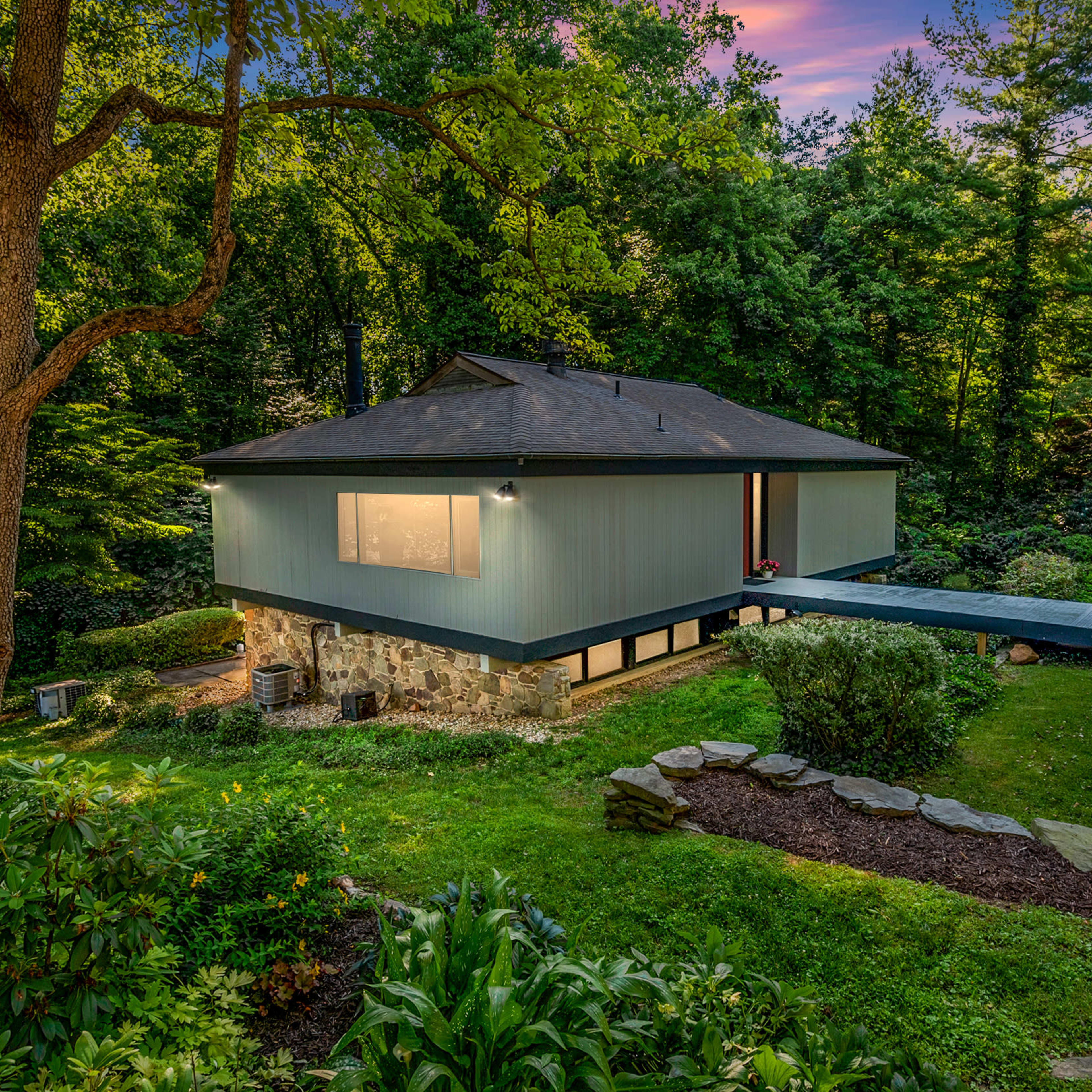 A modern house situated on a hillside, surrounded by lush greenery and a stone pathway leading to the structure.