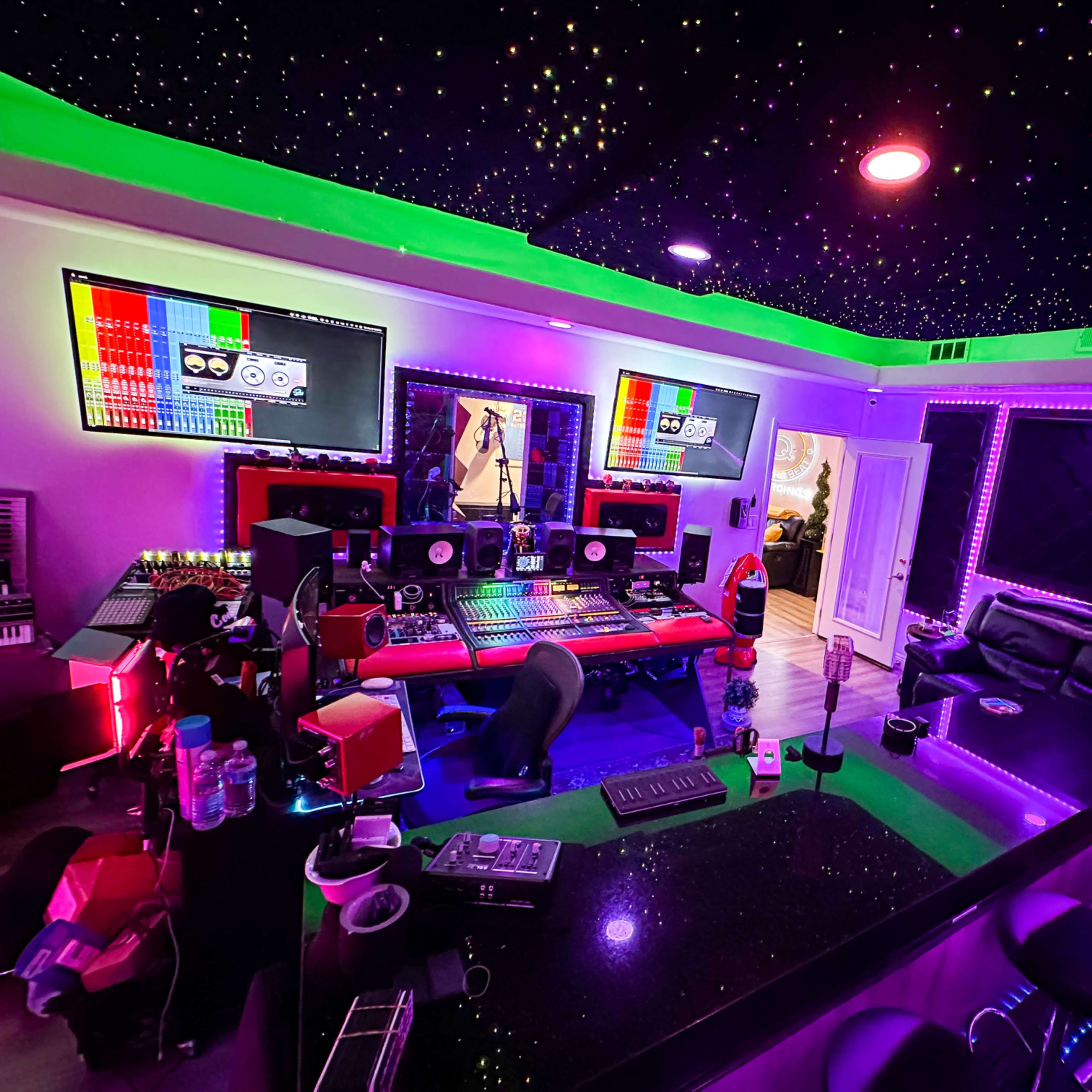 A music studio featuring a black countertop, multiple computer screens, red and green lighting, and a seating area with a black couch.