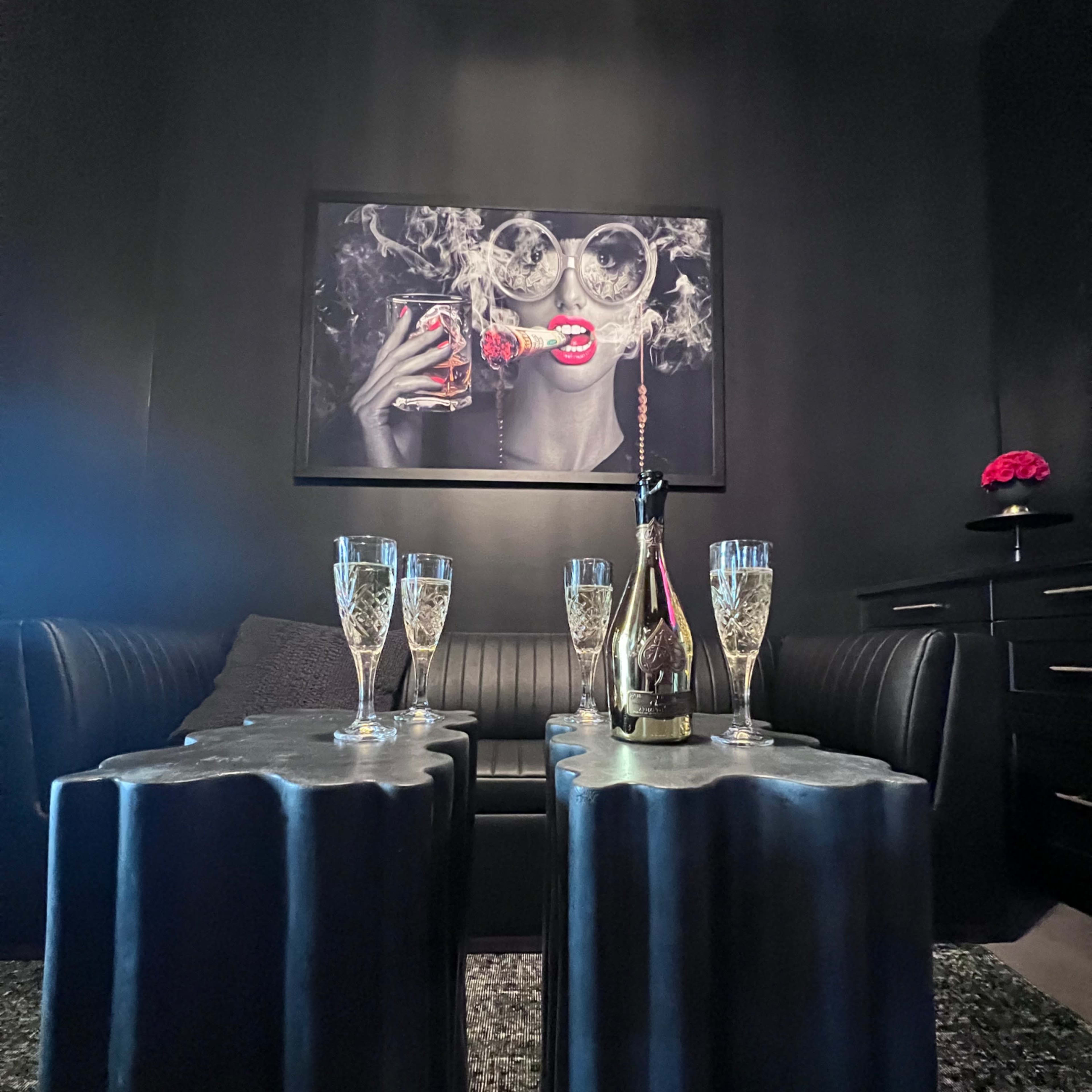 A dimly lit lounge area with black walls, a large framed artwork of a woman, and several champagne flutes arranged on textured black tables.