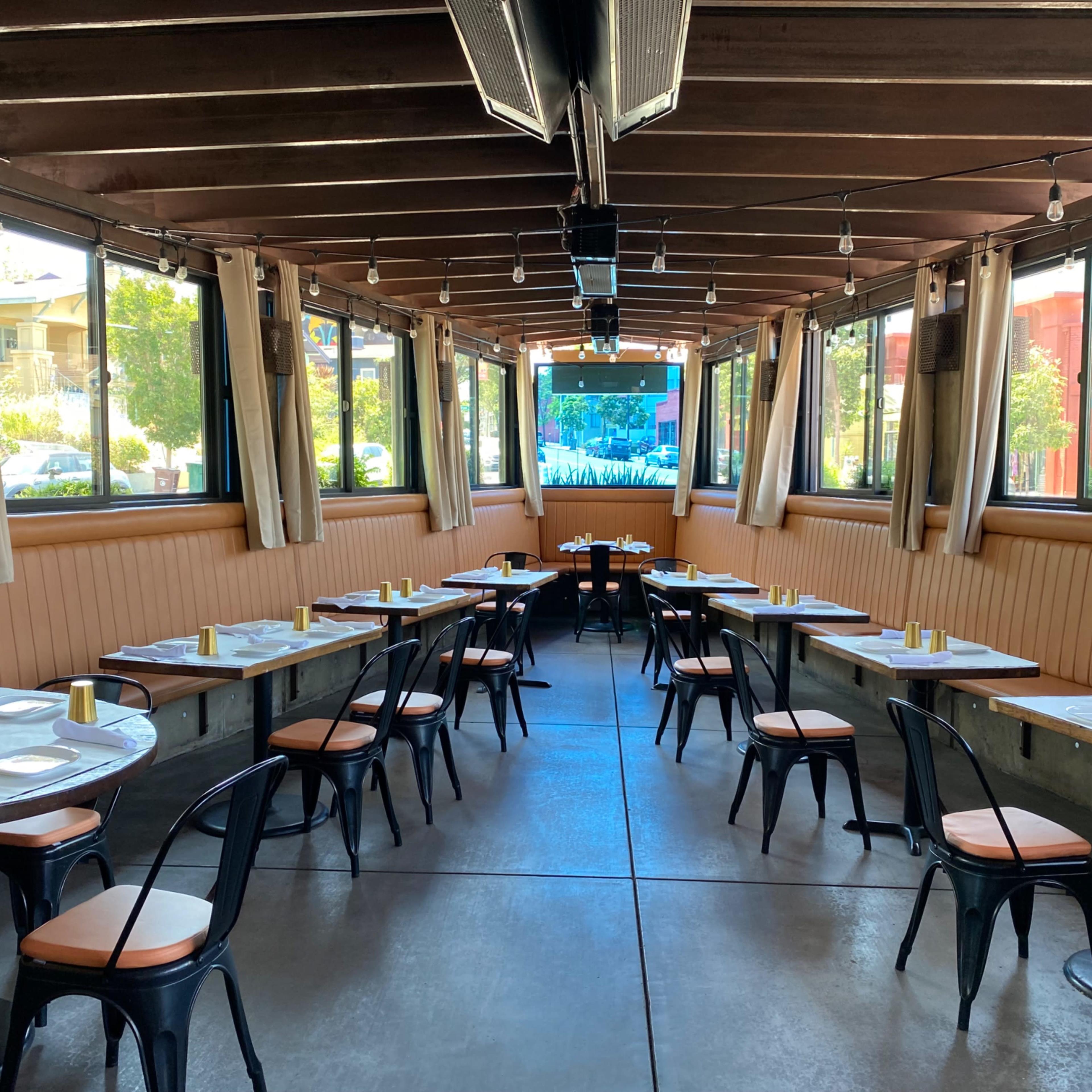 The indoor dining area features several tables with black metal chairs and gold accents, surrounded by large windows letting in natural light.