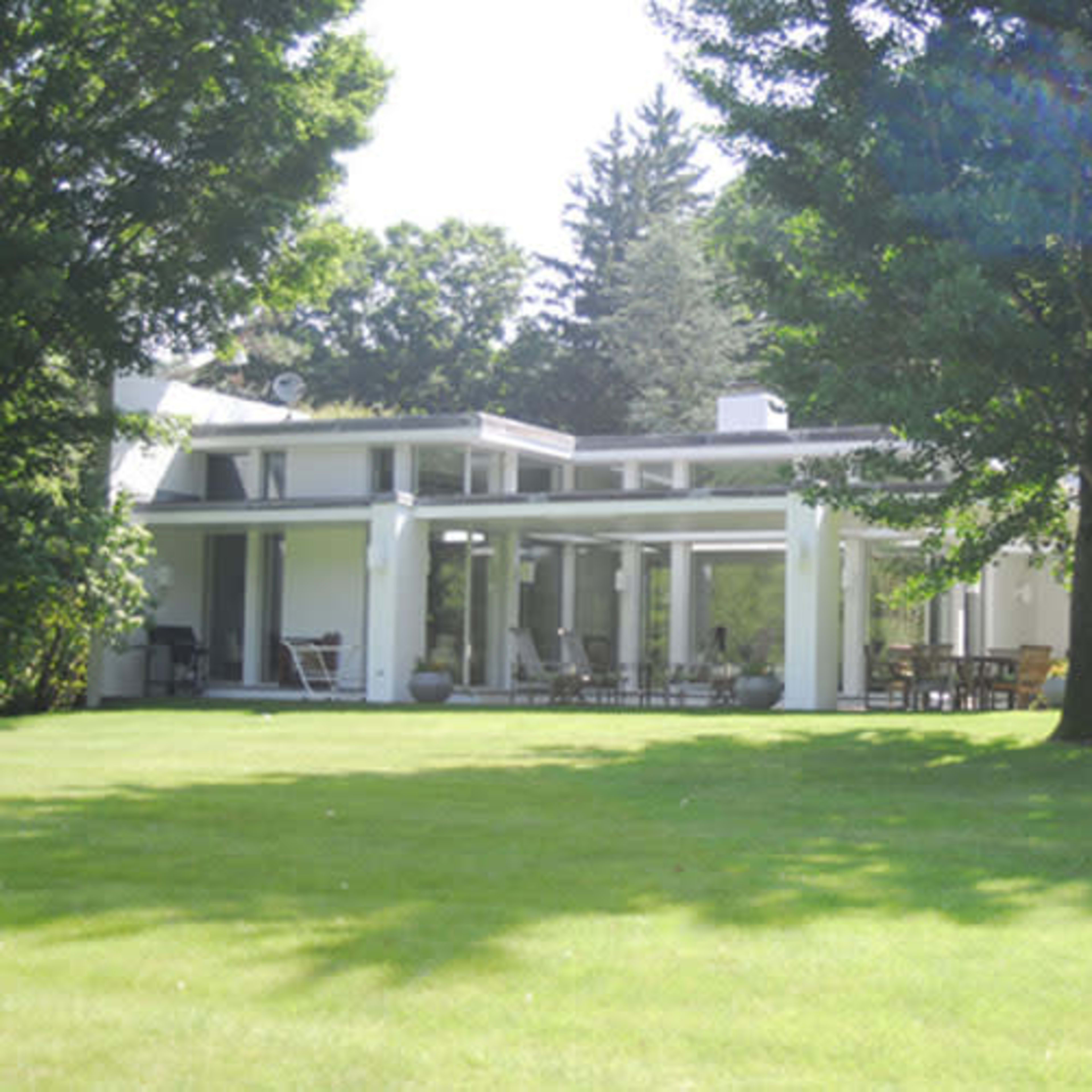 A modern one-story house with large windows and a patio set in a grassy yard surrounded by trees.