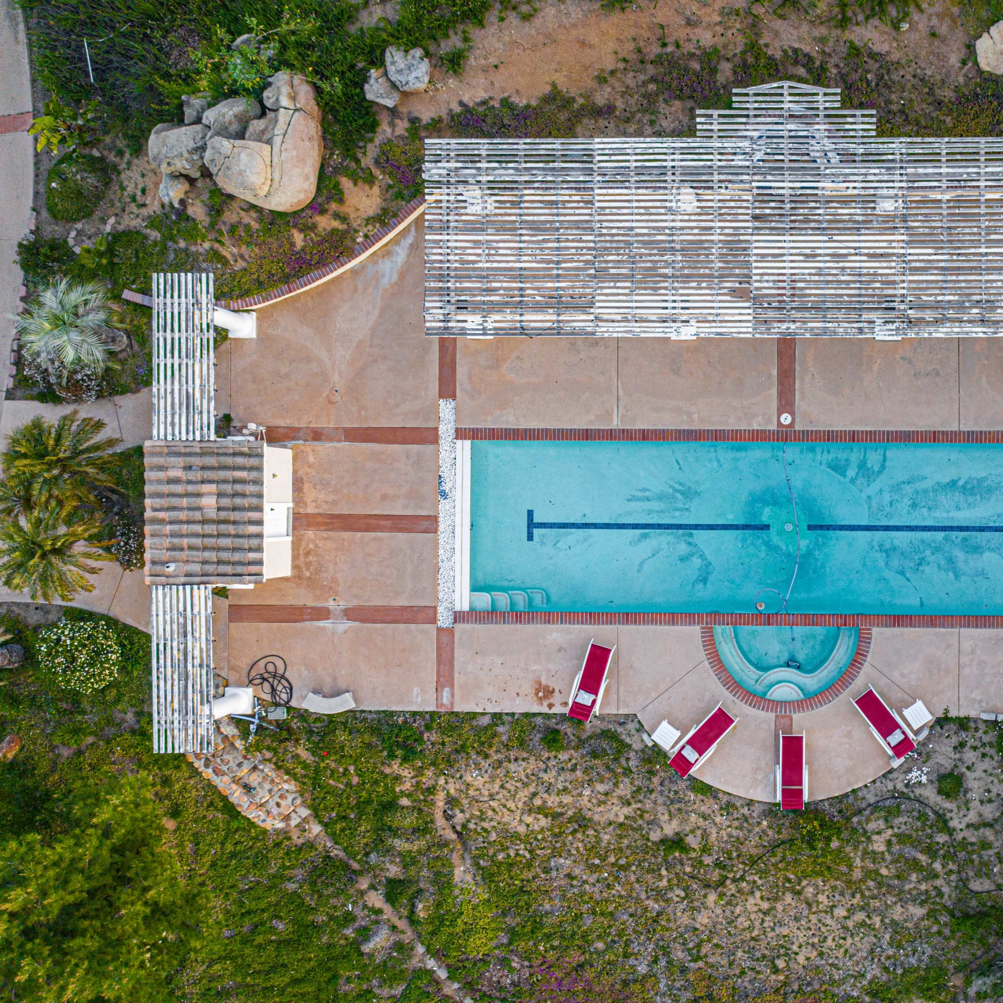 An overhead view of a rectangular swimming pool surrounded by lounge chairs and a pergola on a landscaped area.