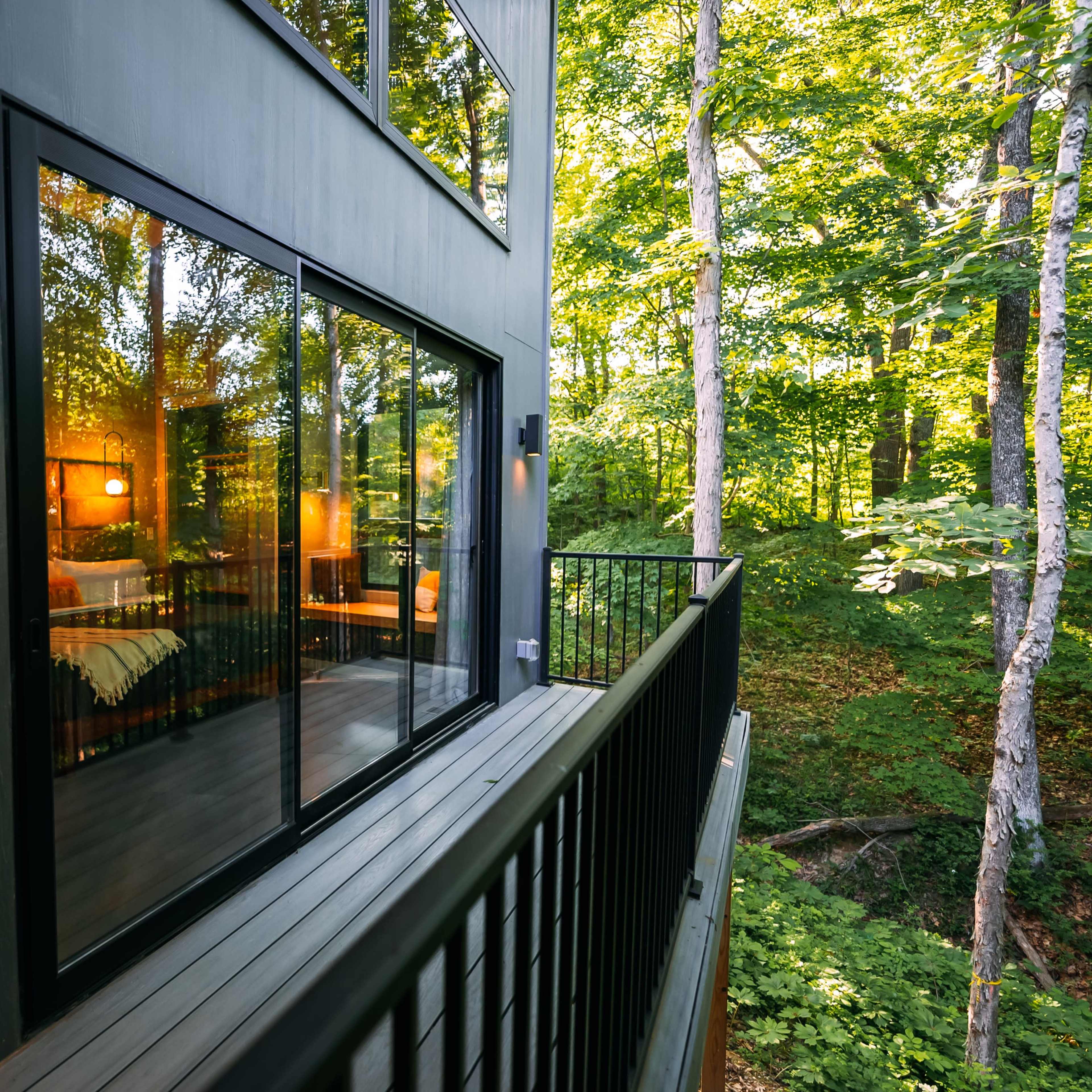 A modern deck of a house surrounded by trees, featuring large glass windows that reflect the greenery outside.