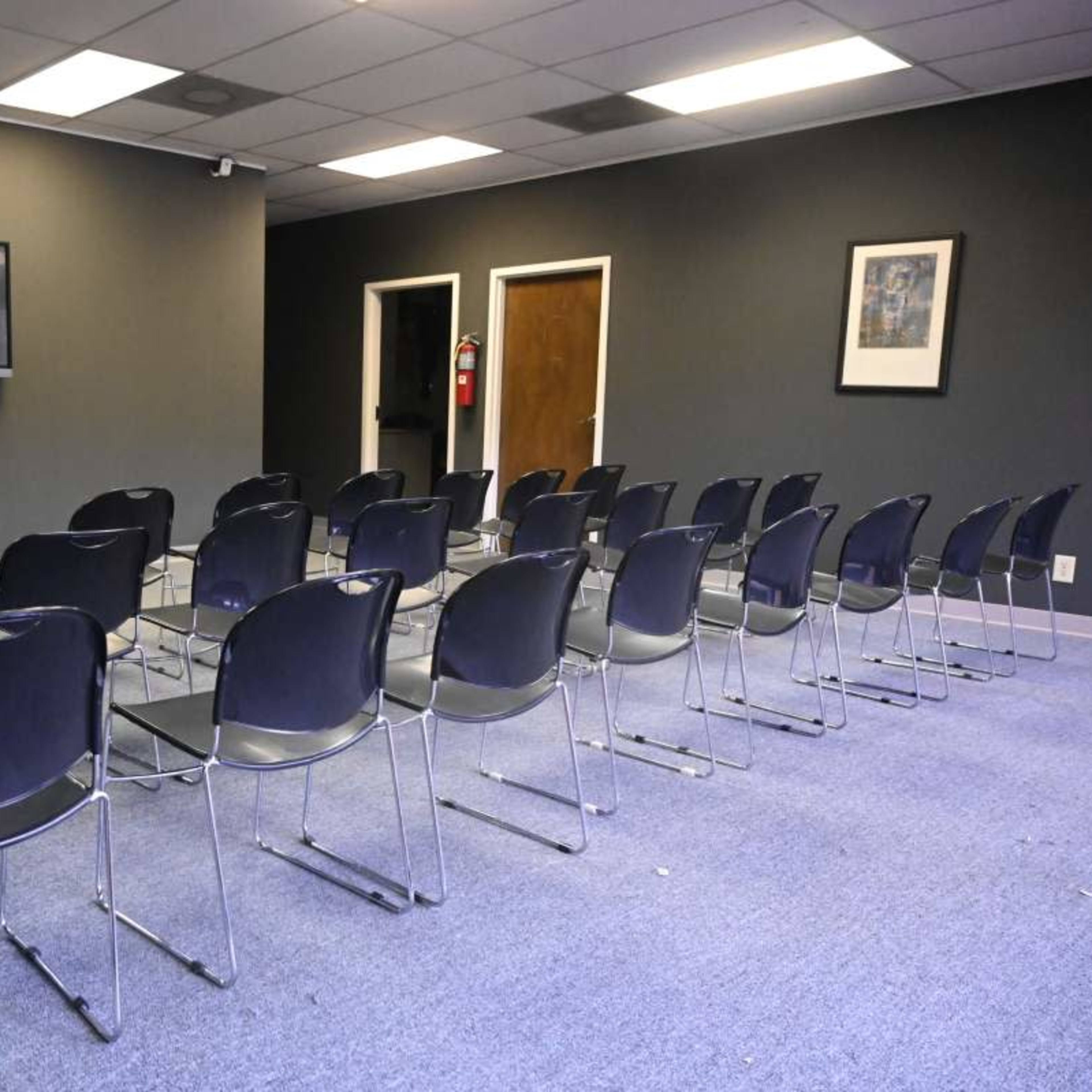 A room with a row of chairs facing a wall-mounted television, with a door and a framed picture visible in the background.