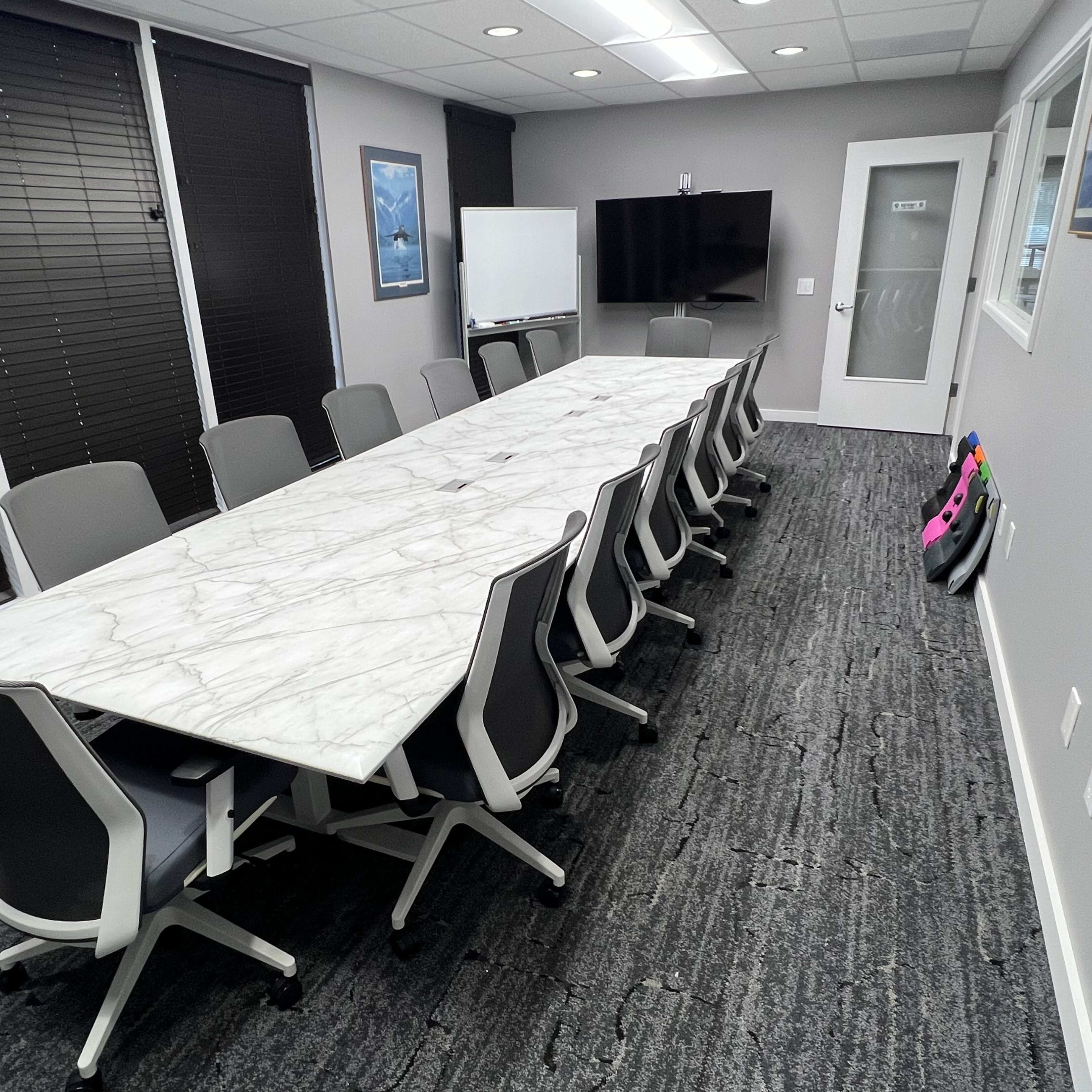 A conference room features a long marble table surrounded by ergonomic chairs, with a large screen and windows dressed with blinds.