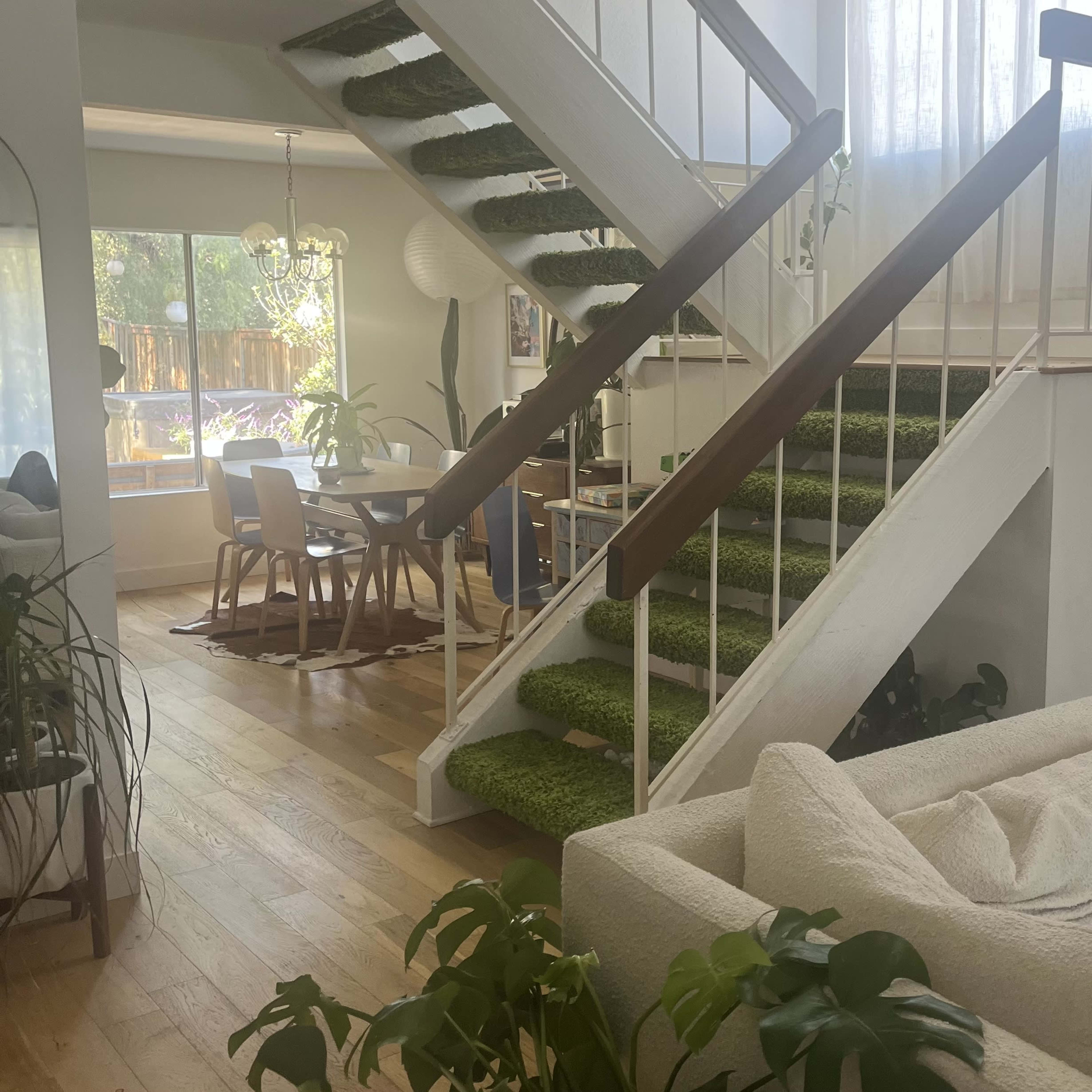 A bright, open living space featuring a staircase with green carpet, wooden flooring, and a cozy seating area surrounded by plants.