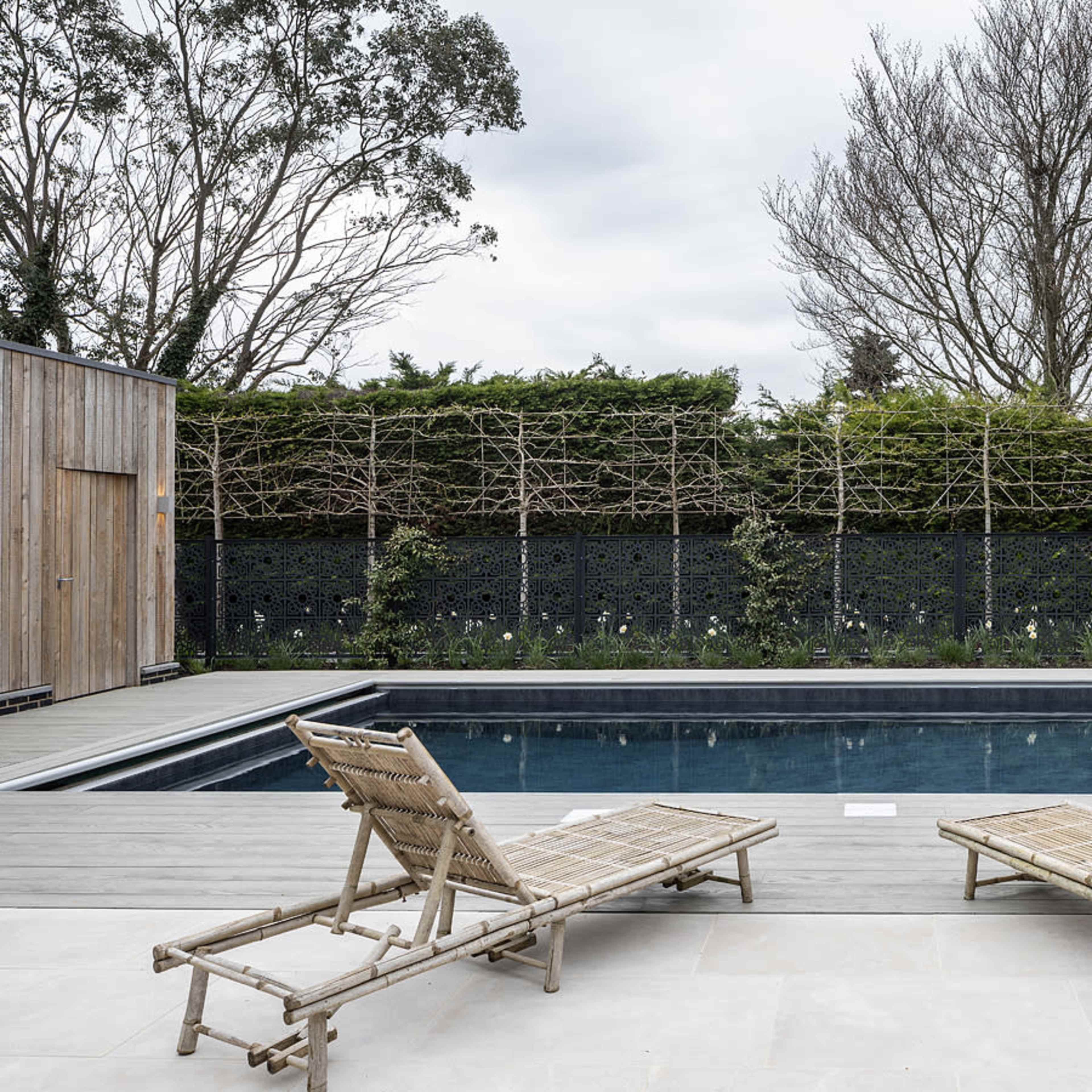 A rectangular swimming pool surrounded by wooden decking and two lounge chairs, with a backdrop of trees and a hedge.