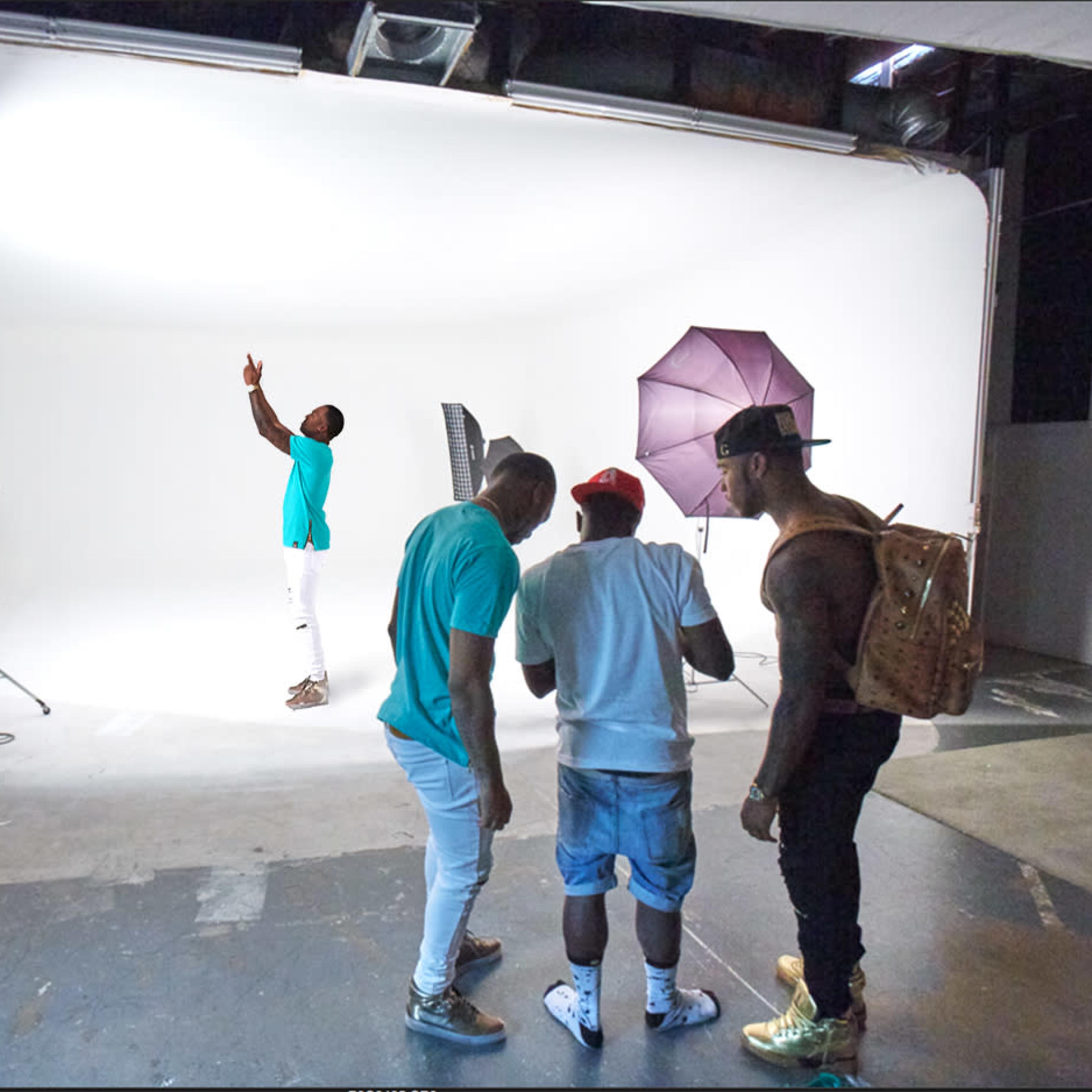 A group of three men is gathered in a photography studio with various lighting equipment and a plain white background.