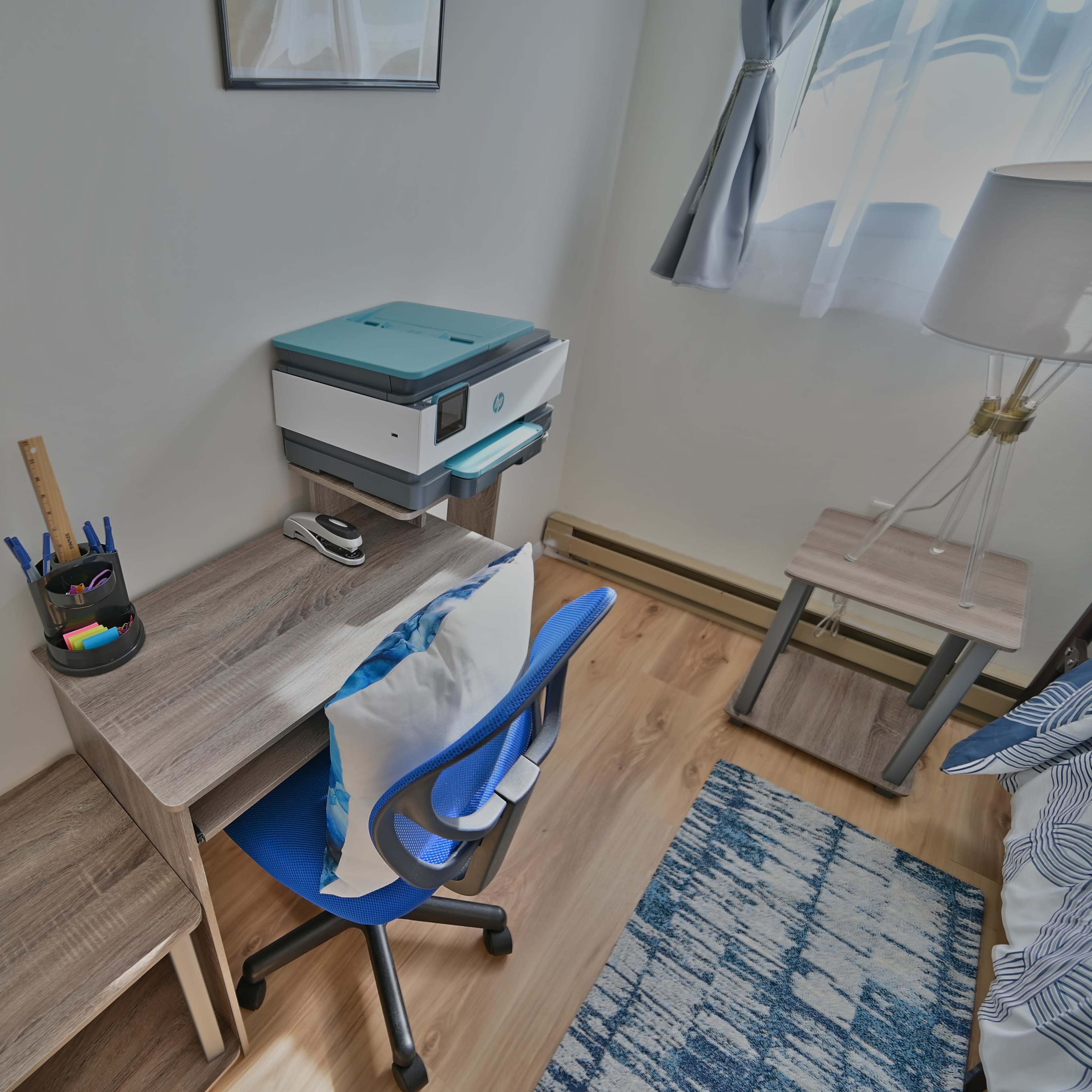 A small workspace is set up in a room with a desk, blue chair, printer, and window curtains.